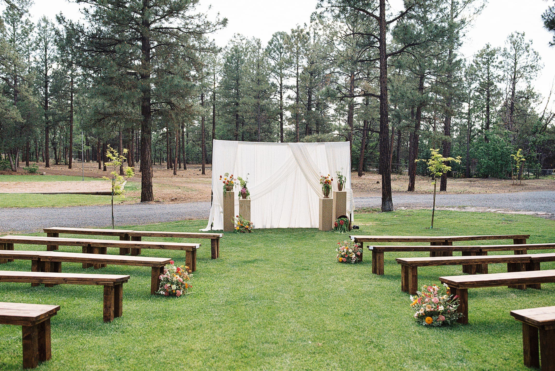 Flowing white draped ceremony backdrop with floral pedestals and wooden benches set on a green lawn in the pines