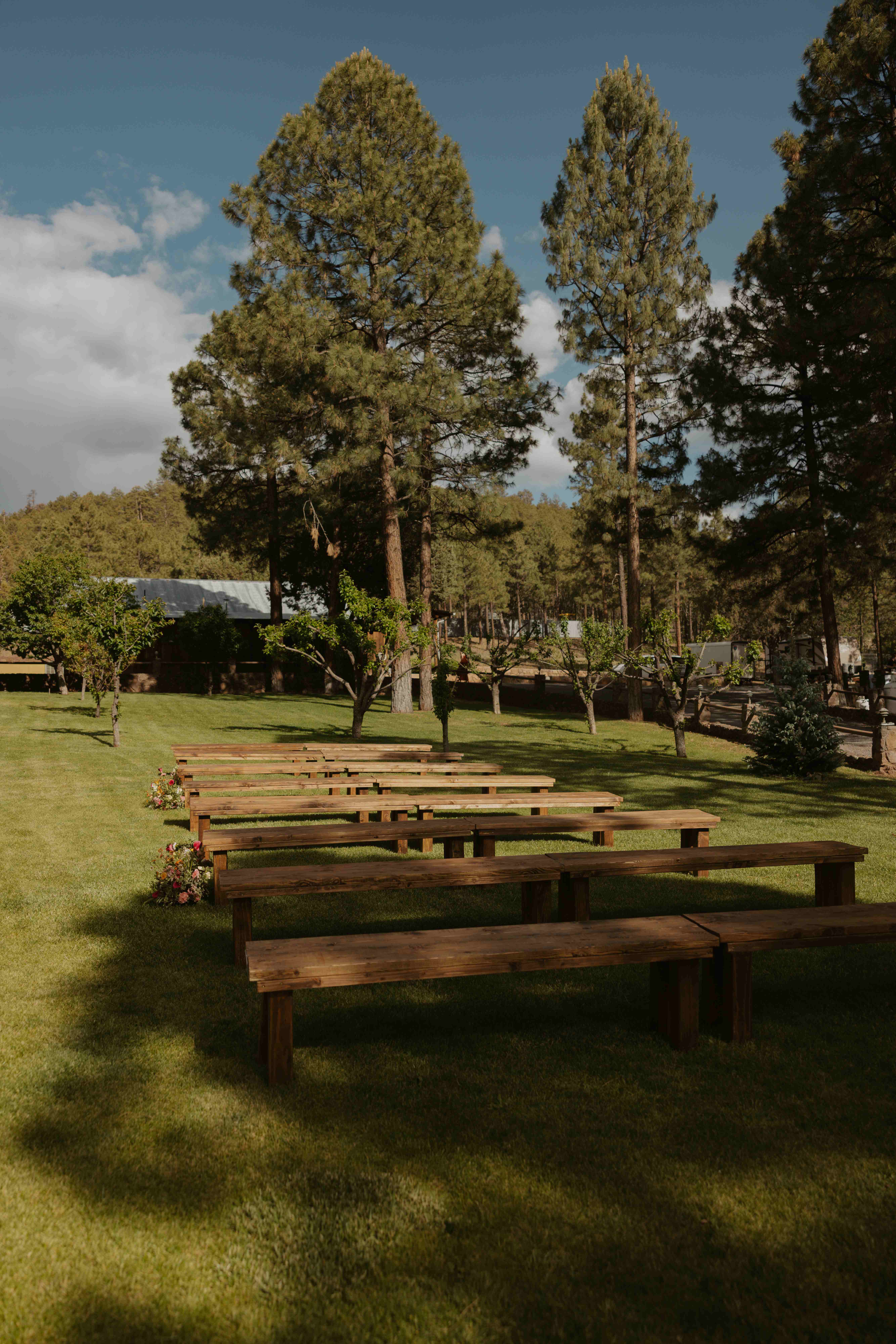 Empty wooden ceremony benches on a sunny lawn with the orchard and ranch buildings in the background