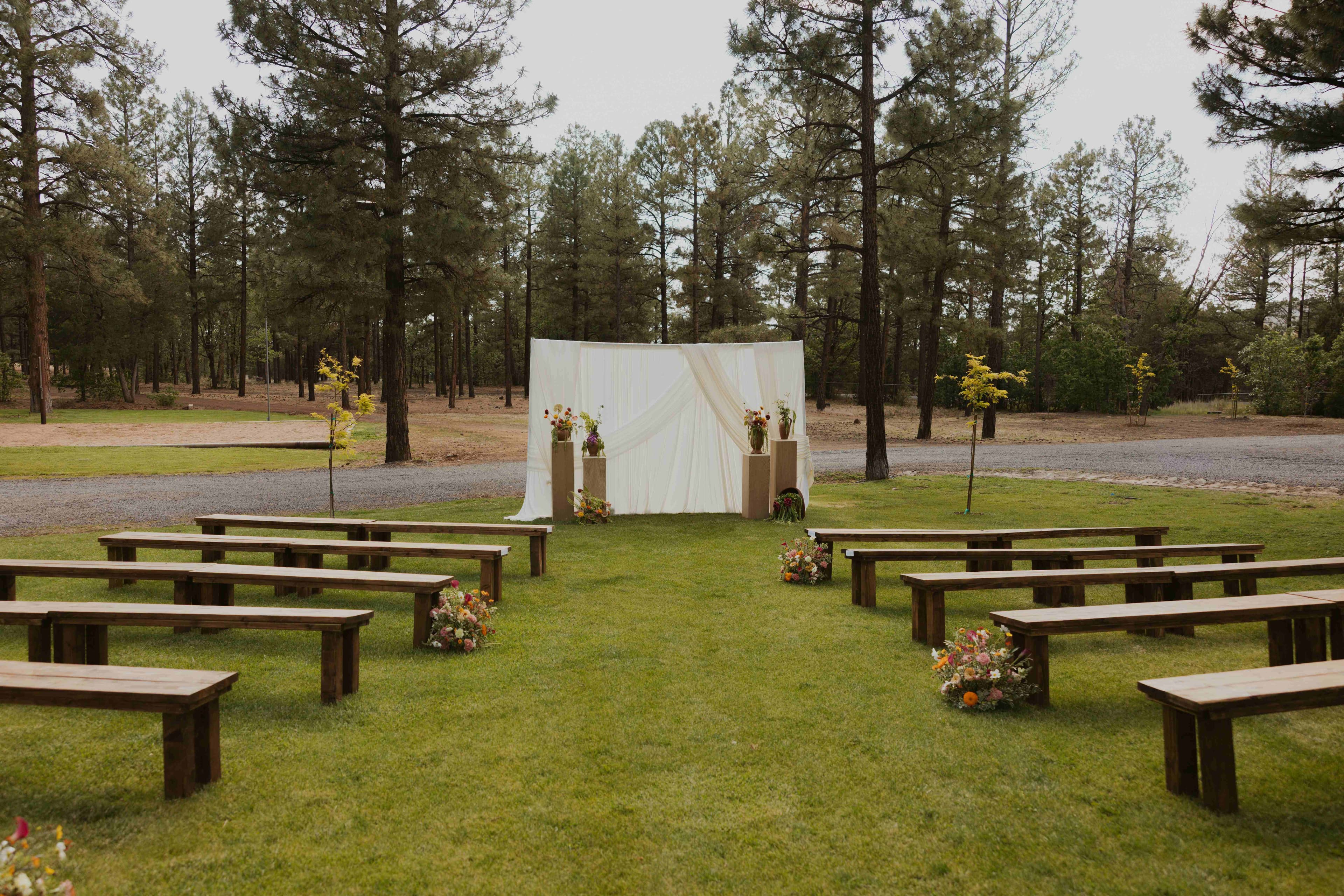 Bride in white beside bridesmaids in warm-toned dresses during a ceremony with a log cabin in the background