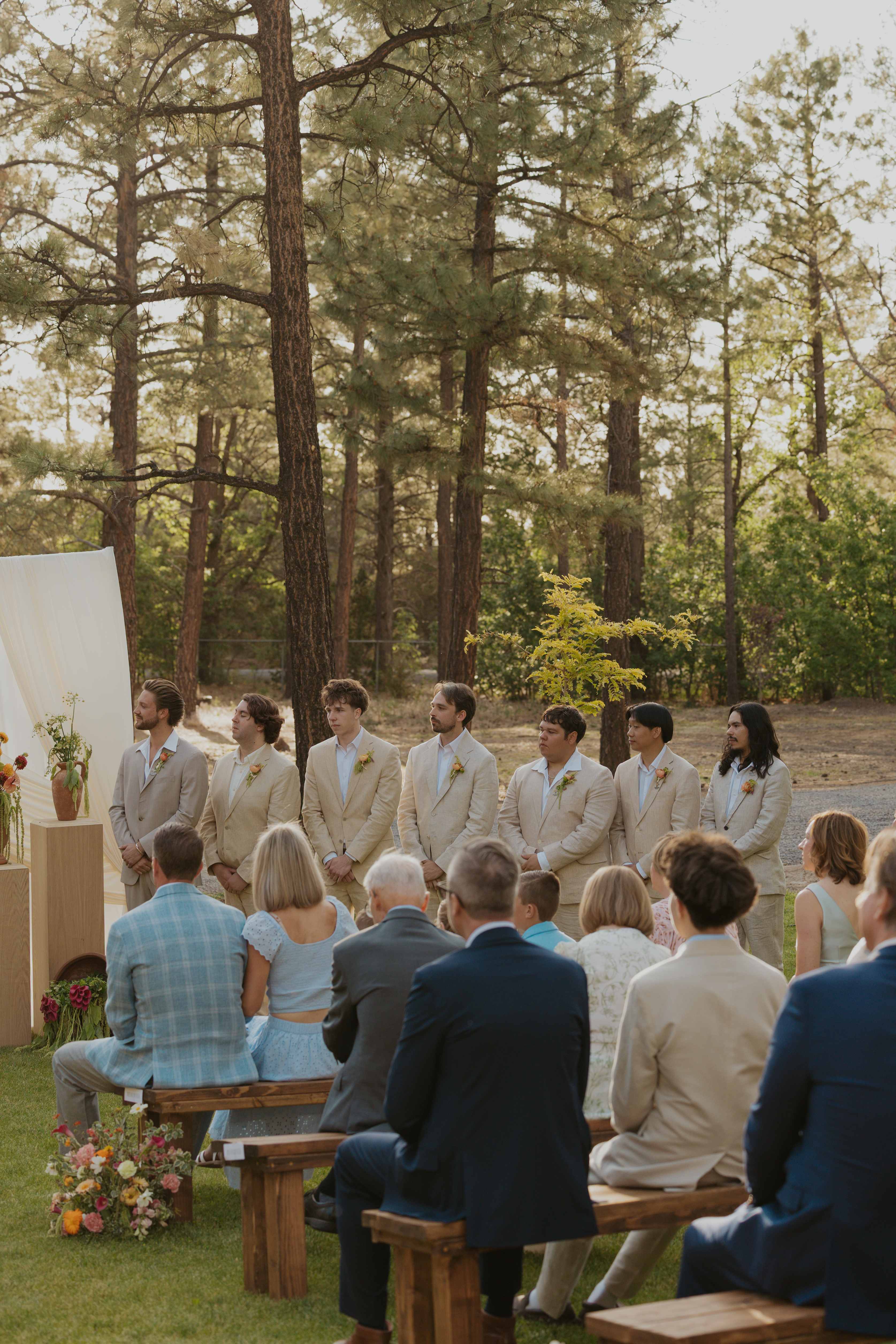 Groom and groomsmen in tan linen suits standing at the front of an outdoor forest ceremony