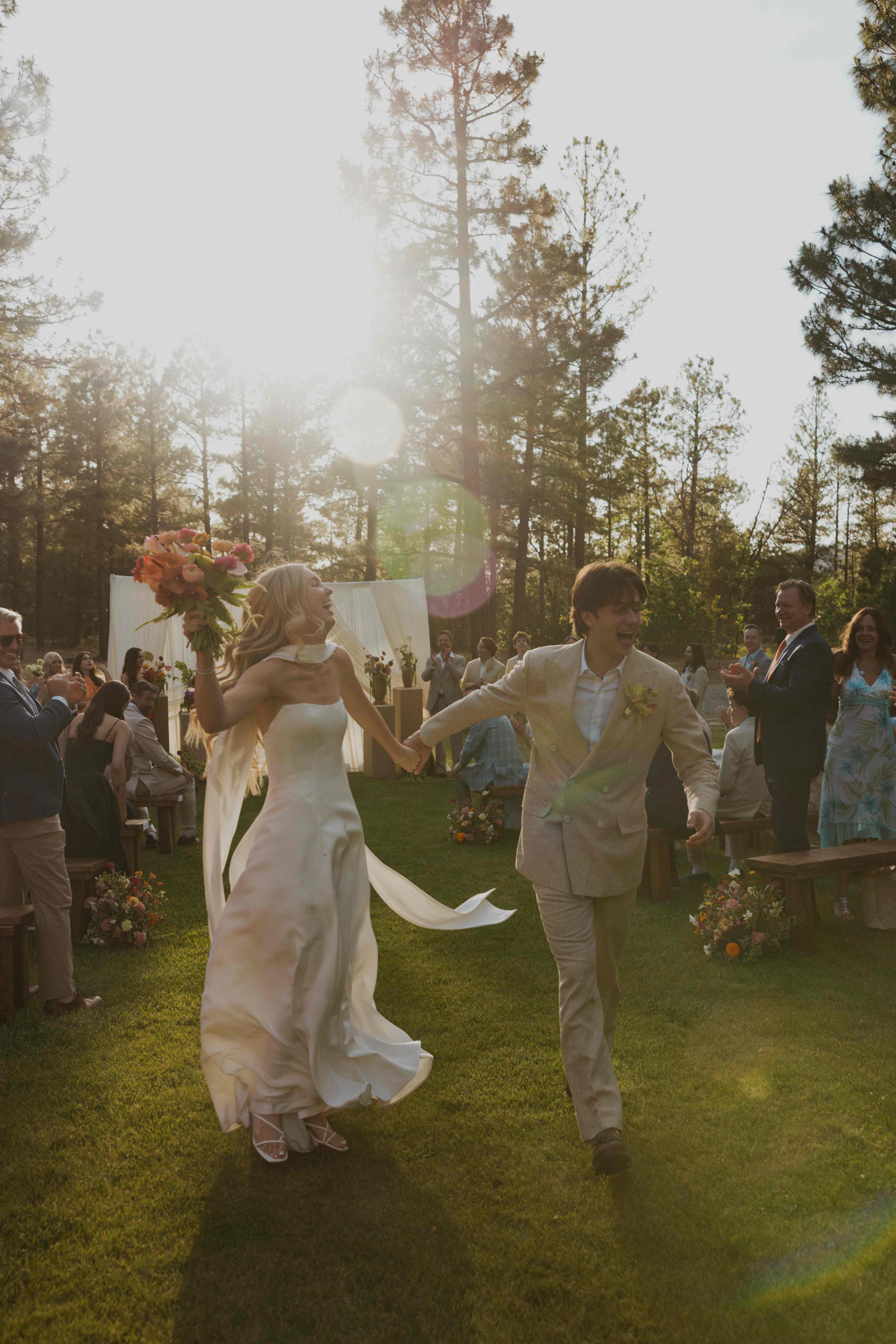 Newlyweds walking back up the aisle hand in hand, the bride holding a colorful bouquet, sun flaring behind them