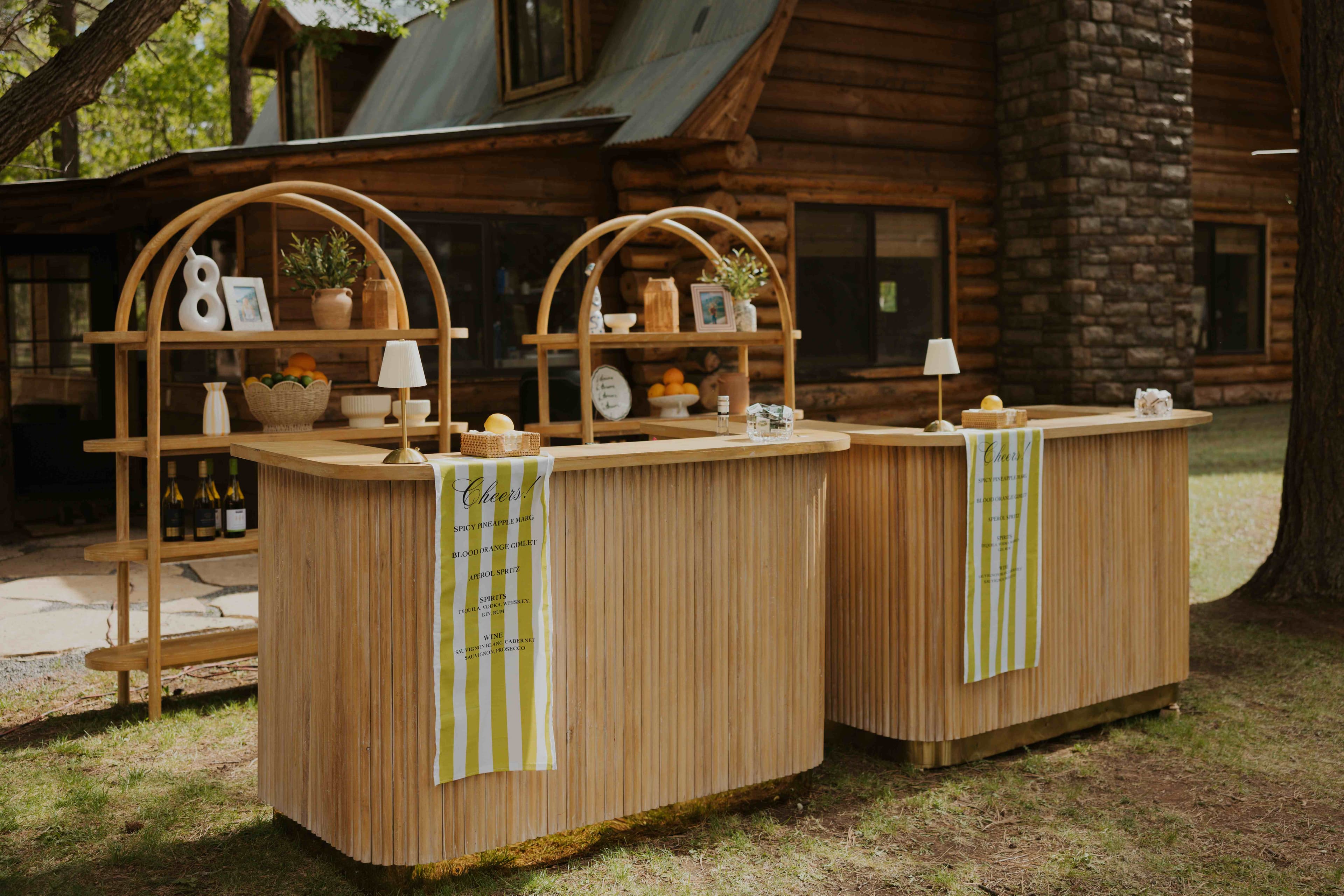 The two curved wood bar stations styled with fruit, greenery, and striped Cheers! menu banners in front of the cabin