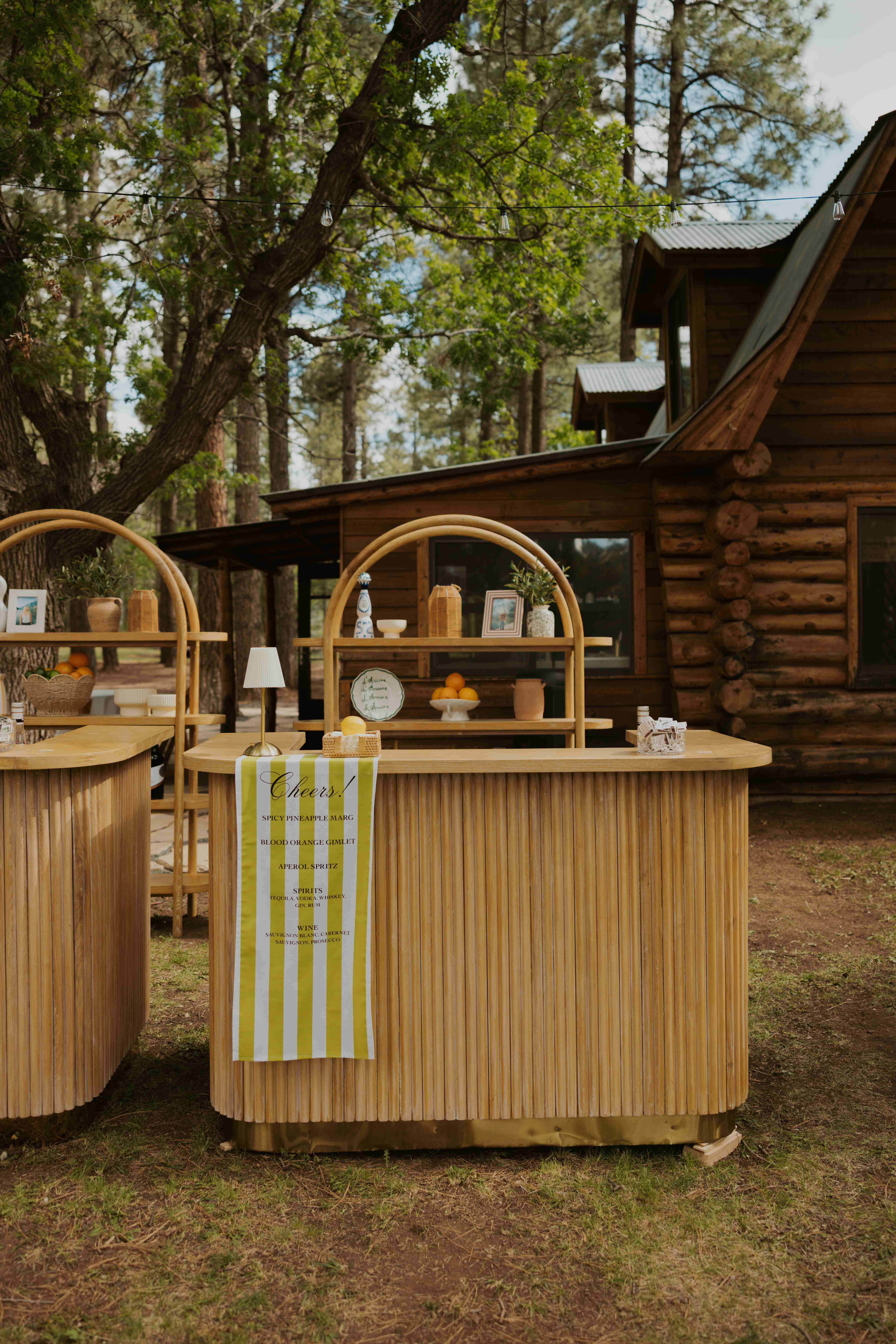 Two curved fluted-wood bar stations with arched rattan shelves and chartreuse-striped Cheers! menu banners beside a log cabin in the pines
