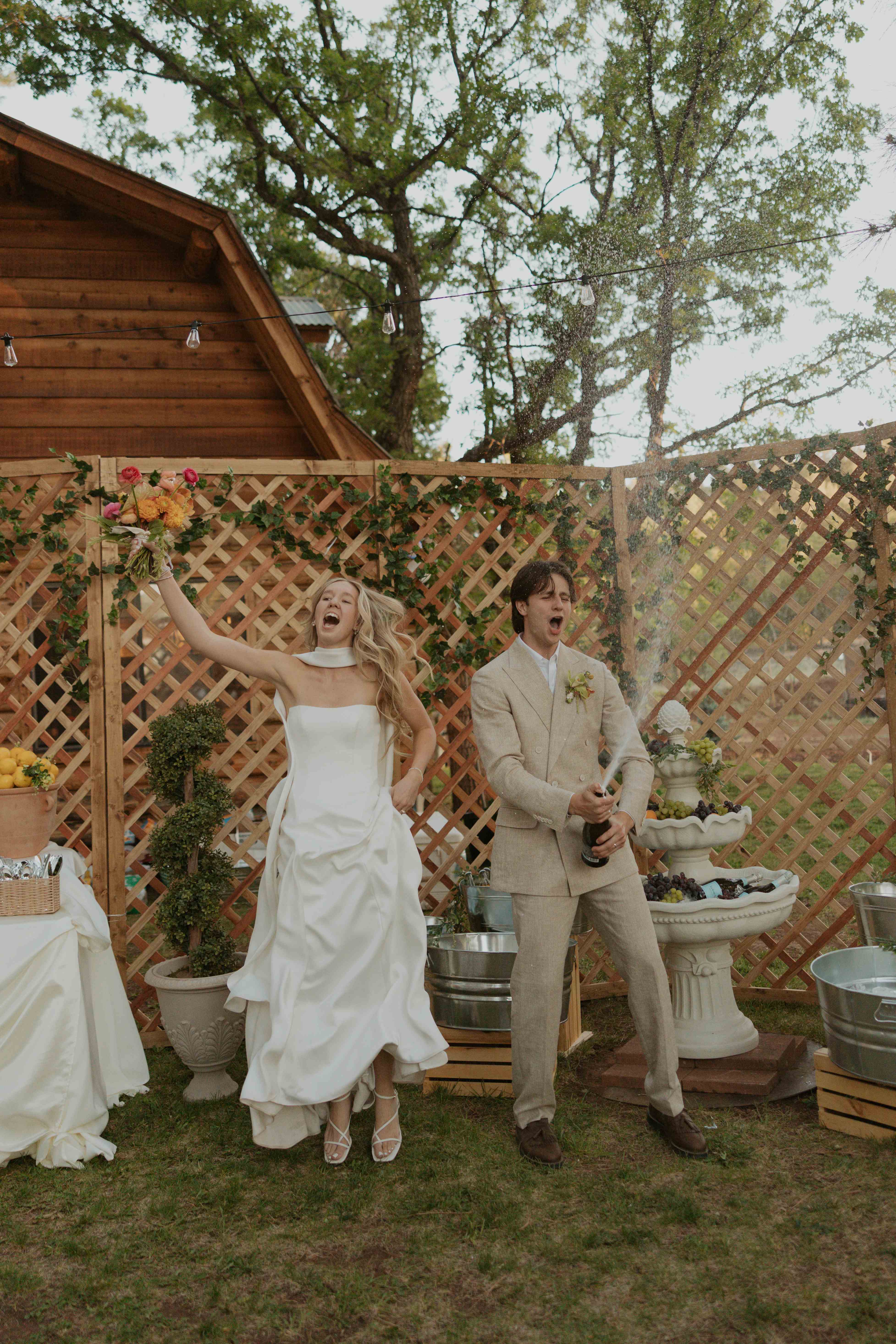 Bride cheering with her bouquet raised while the groom sprays champagne against a lattice fence with lemons and a tiered fountain