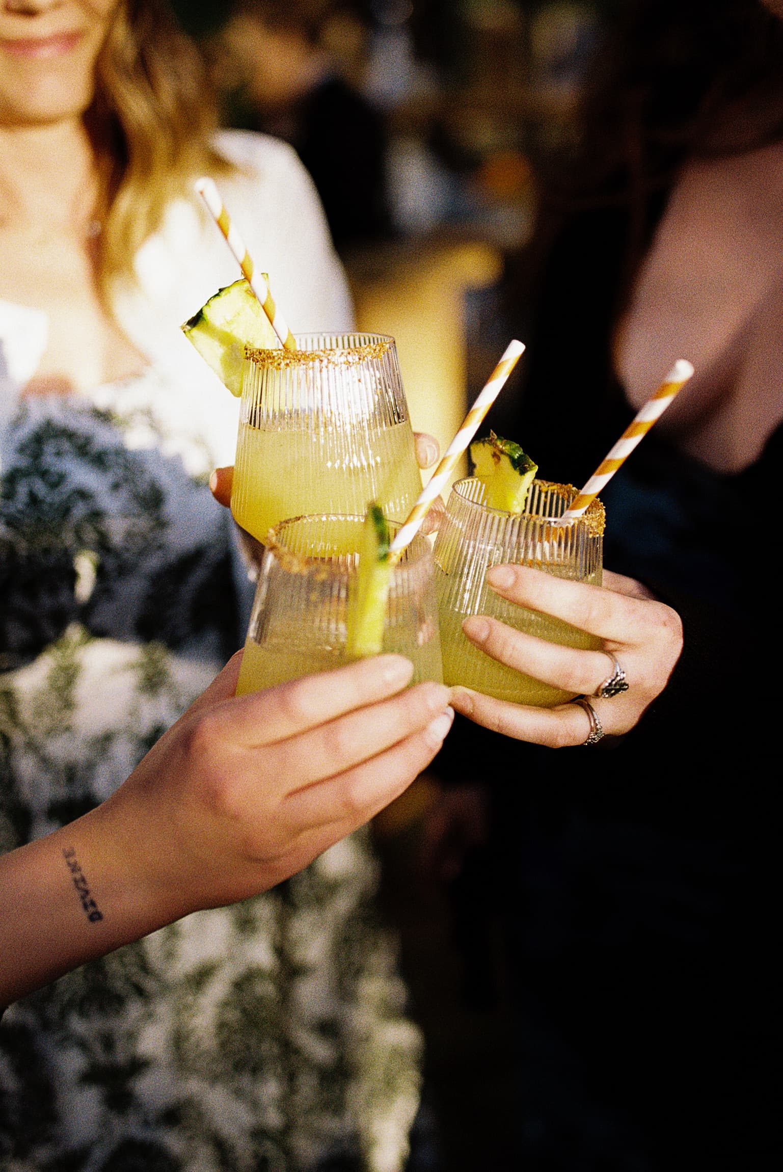 Close-up of three hands toasting pineapple-garnished cocktails with striped paper straws