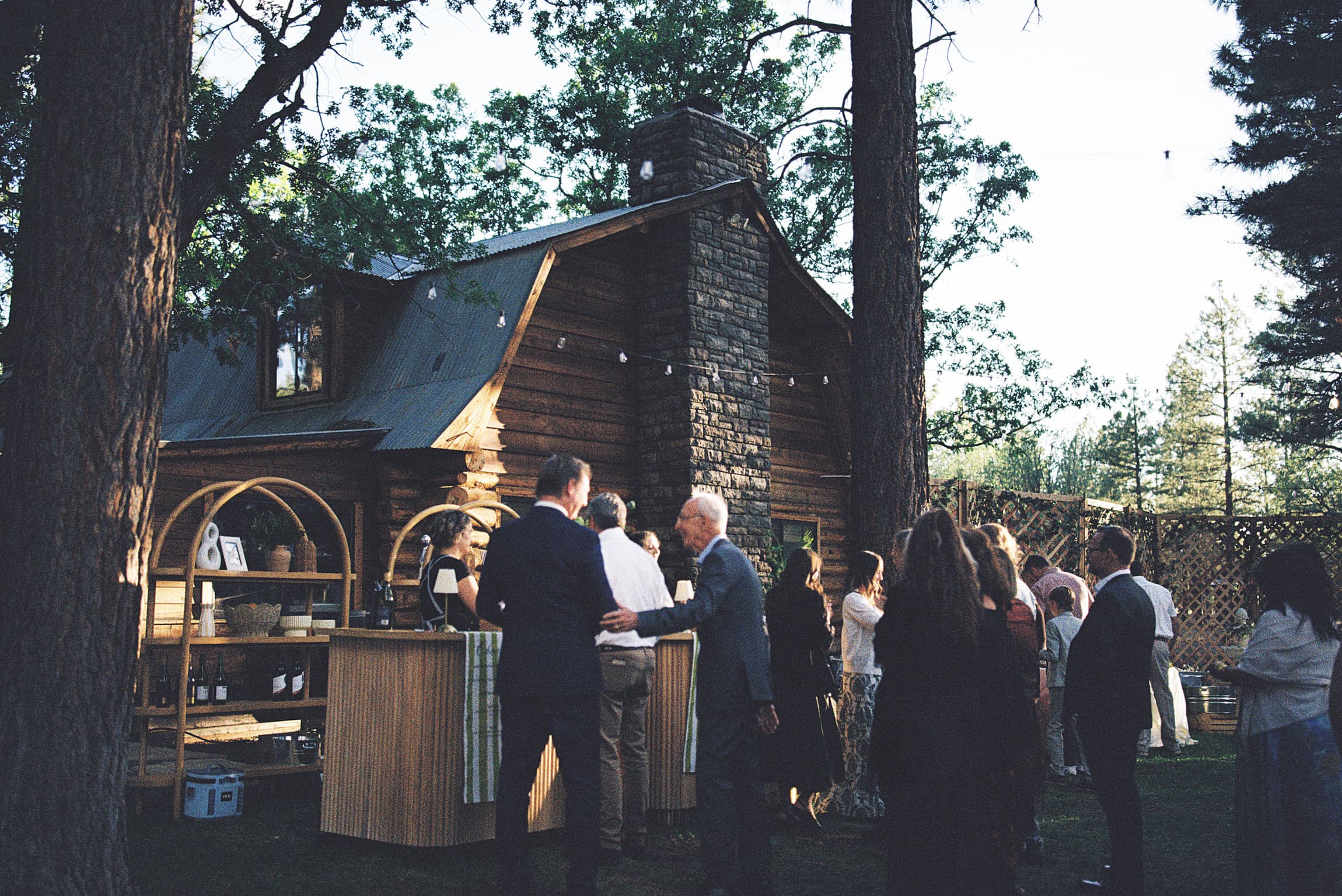 Guests mingling at the wooden bar beside the stone-chimney log cabin at golden dusk