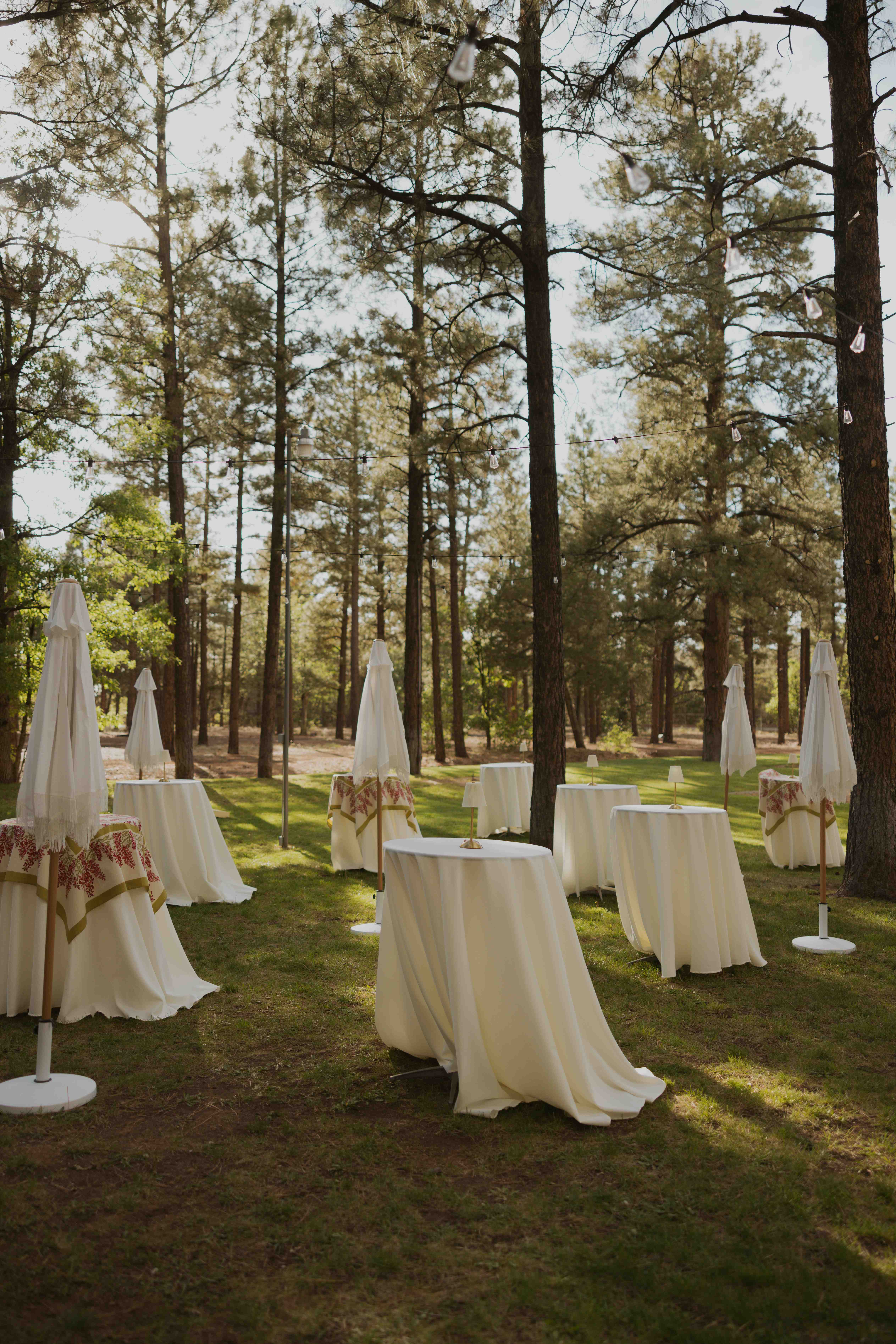 White-linen cocktail tables with closed fringed umbrellas and small lamps set across a grassy clearing under tall Ponderosa pines