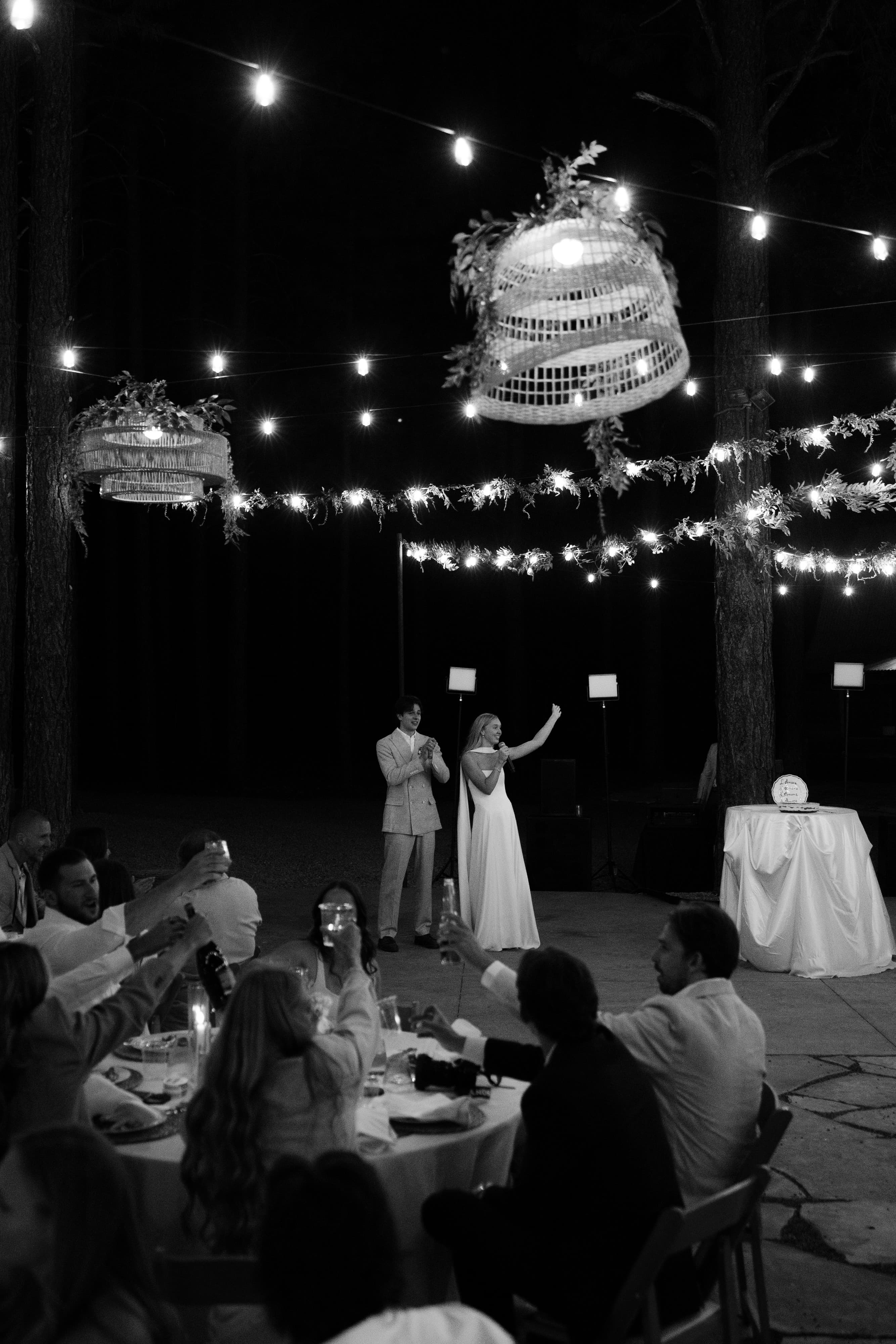 Black-and-white nighttime photo of the couple giving a speech with a microphone under woven lanterns as guests toast