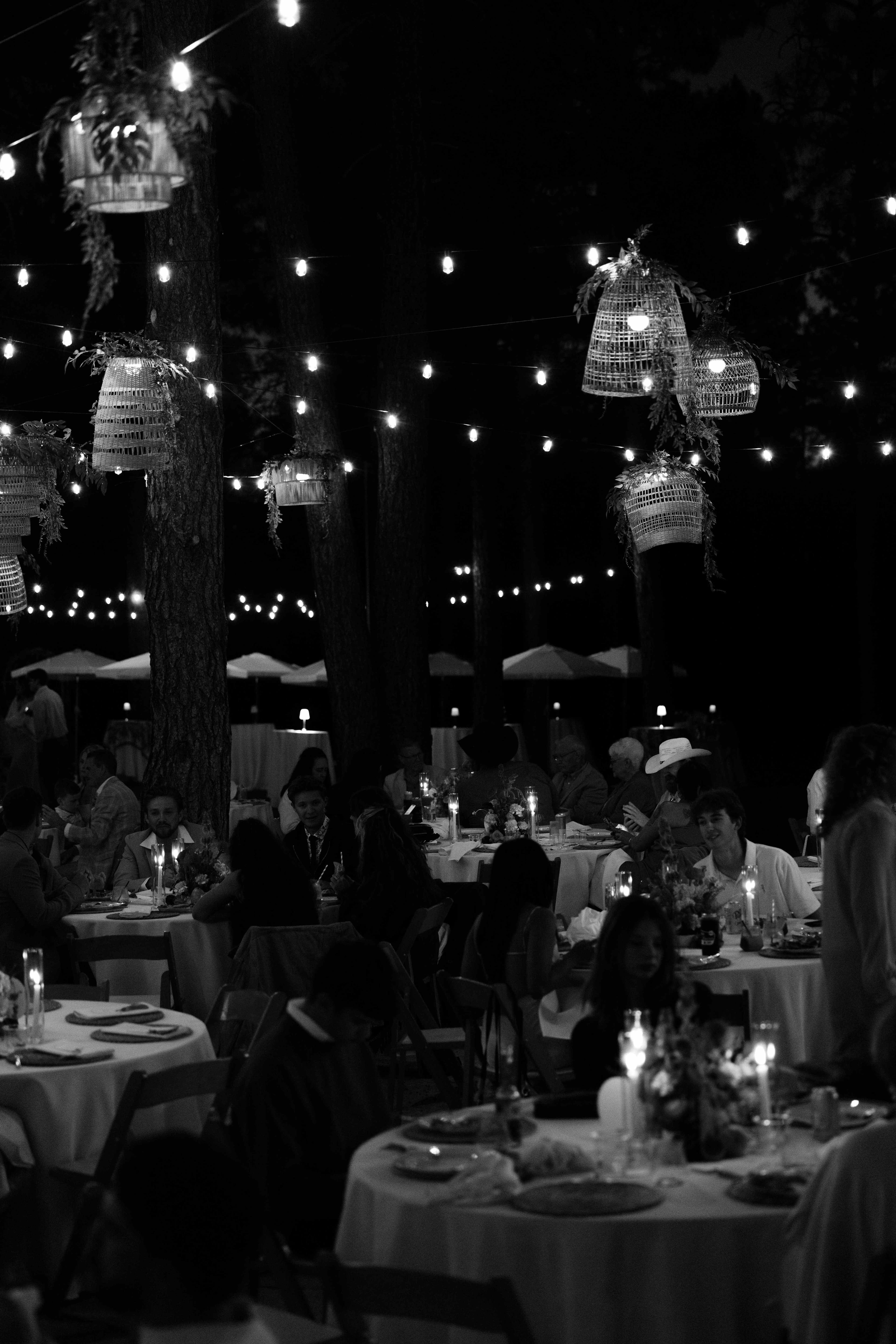 Black-and-white nighttime reception with glowing woven rattan lanterns and string lights over round candlelit tables of seated guests