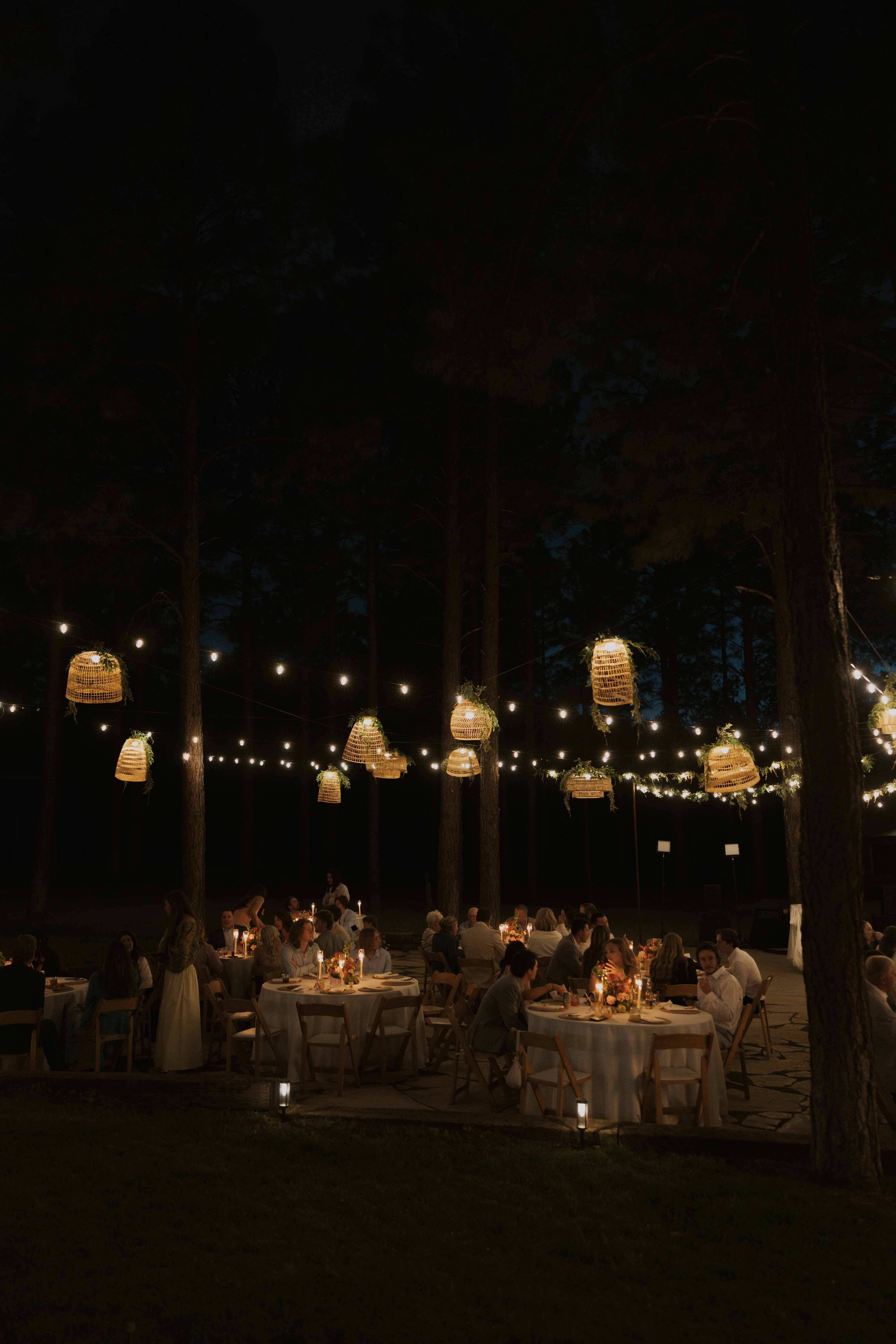Moody wide nighttime view of glowing woven lanterns and string lights over dim candlelit reception tables in the forest