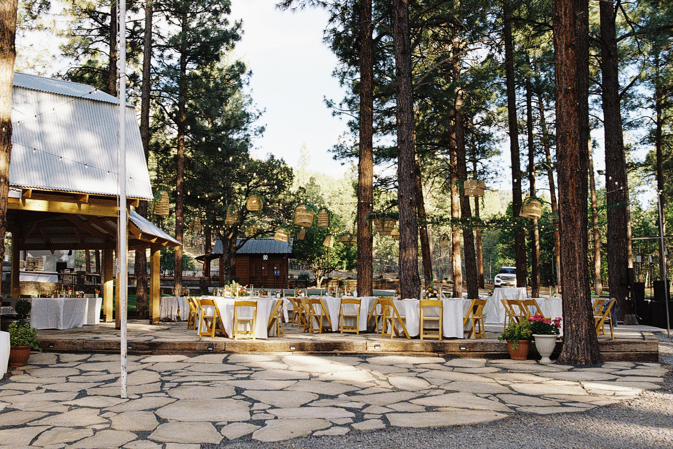 Long reception tables with white linens and yellow folding chairs set beside the ramada among the tall pines