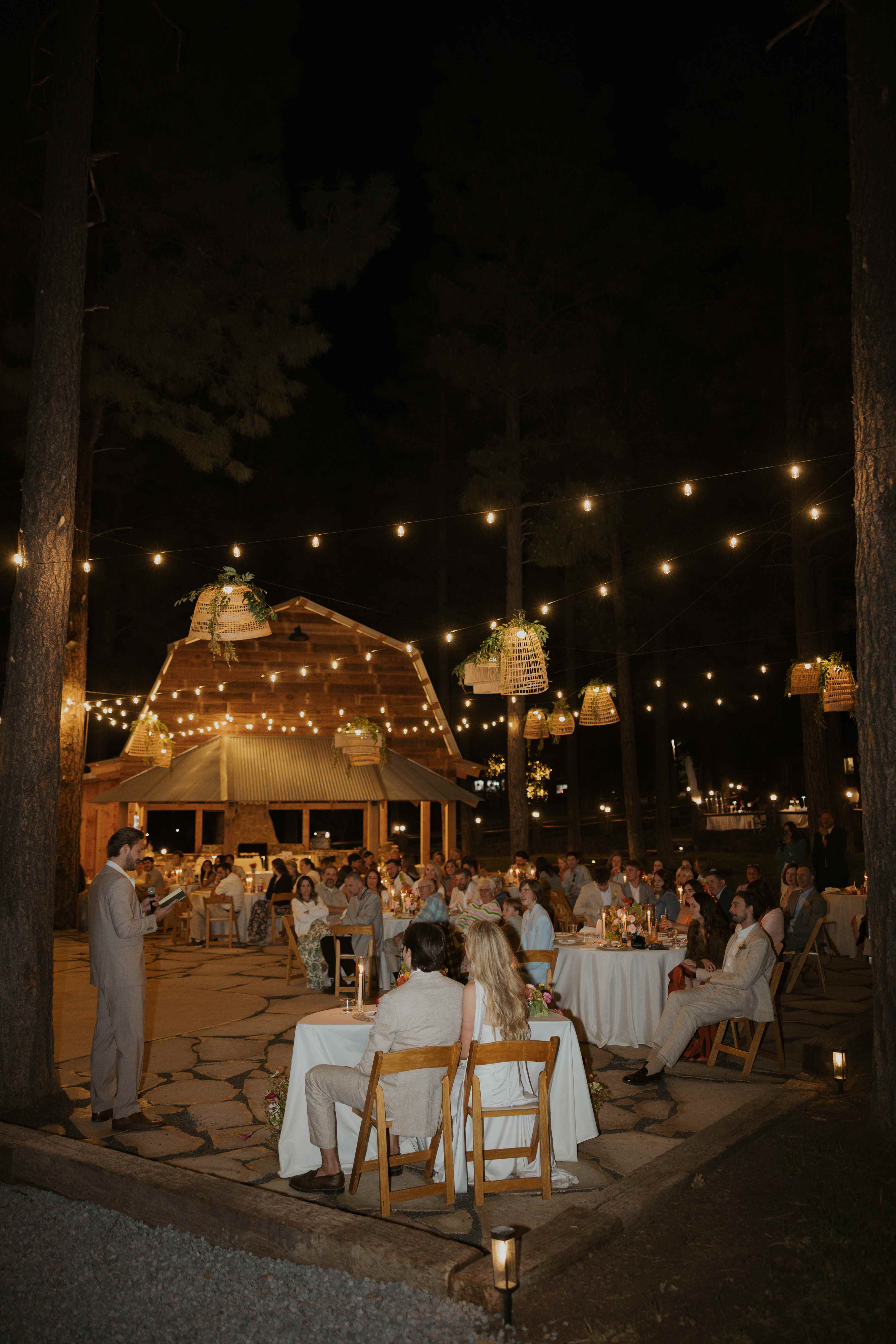 Groom giving a speech to guests at candlelit reception tables at night under woven lanterns, the barn glowing behind