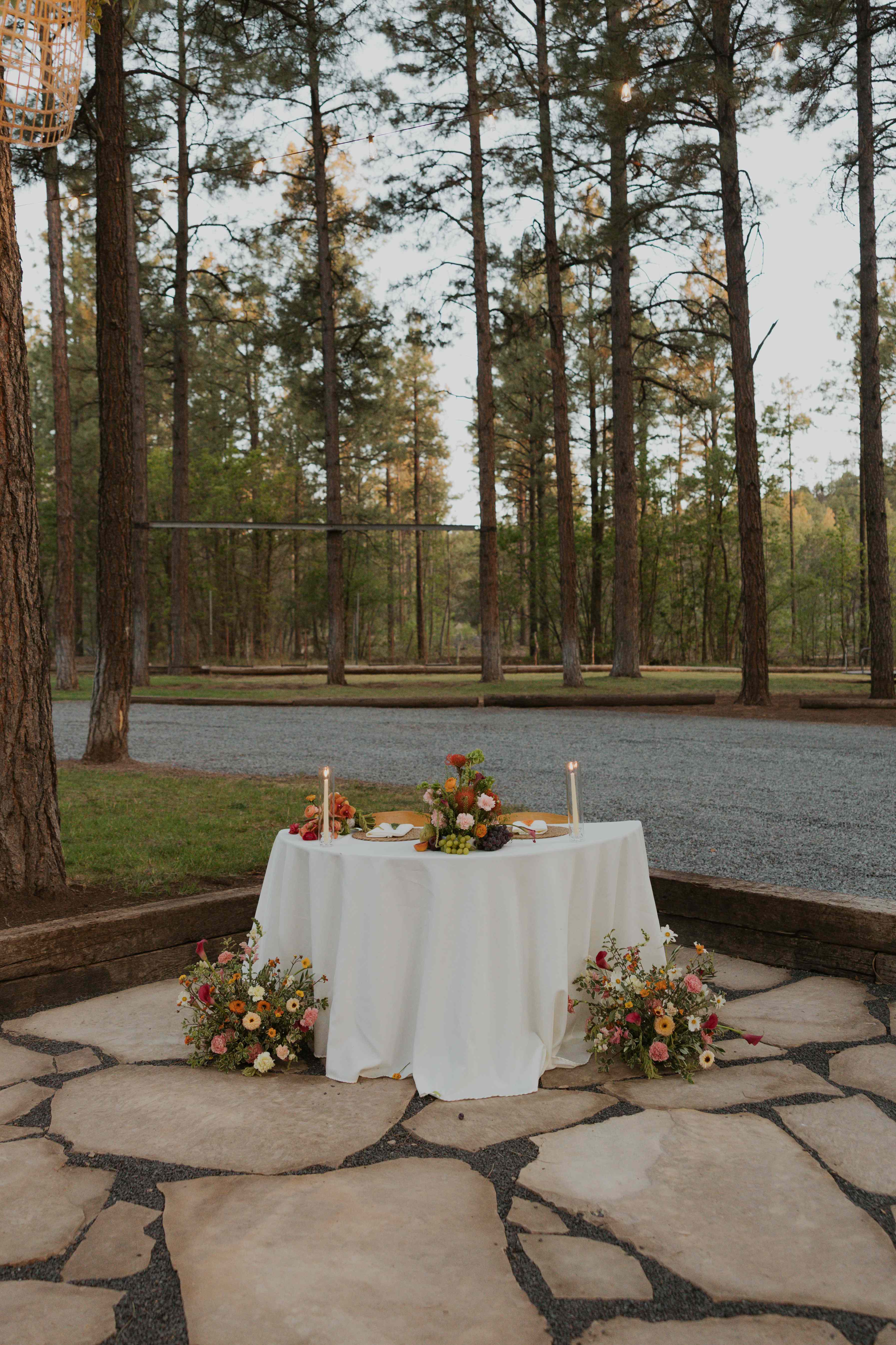 Sweetheart table draped in white linen and flanked by wildflower arrangements at dusk on the flagstone patio in the pines
