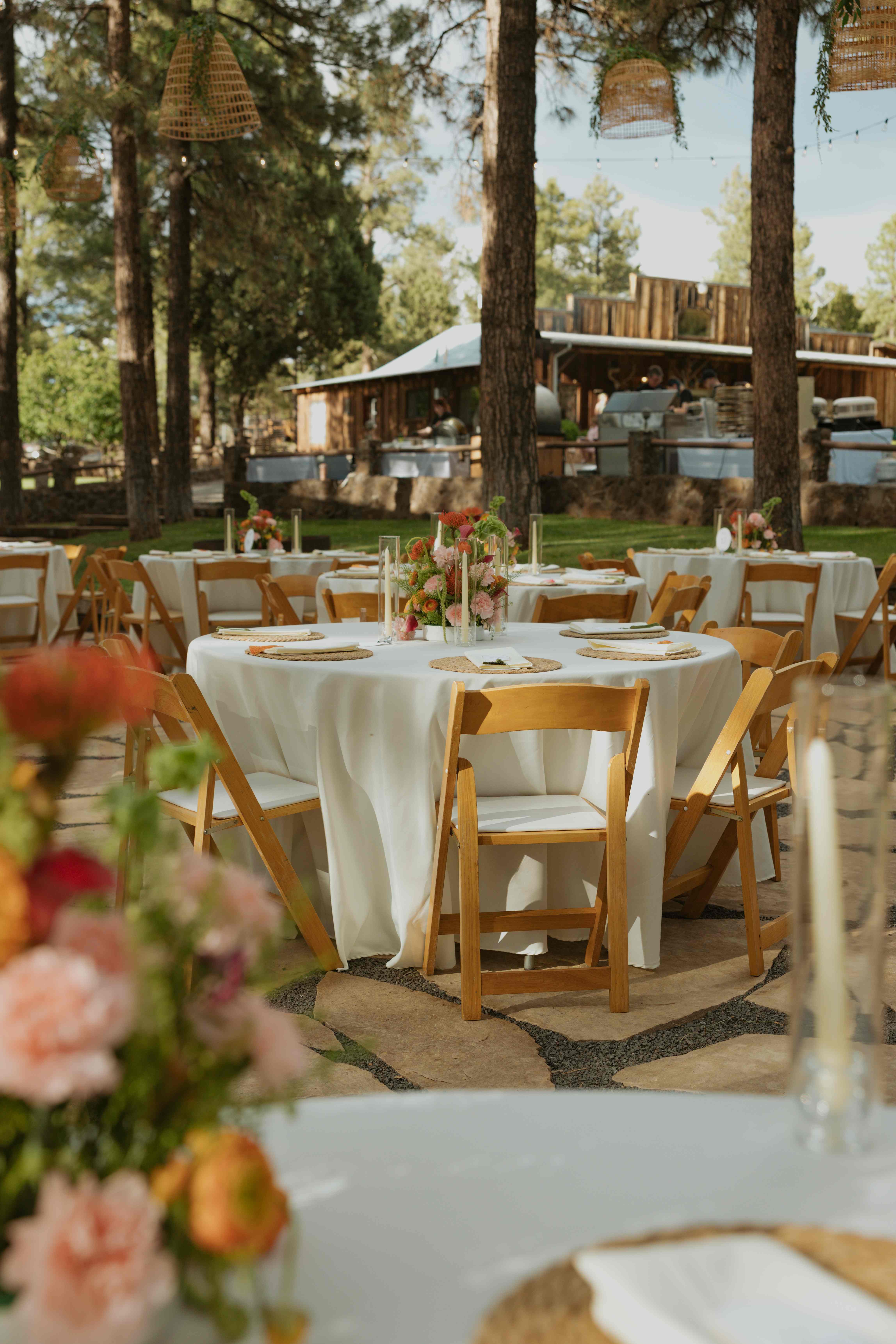 Daytime reception with round tables, floral centerpieces, and wooden chairs, a catering station by the cabin behind