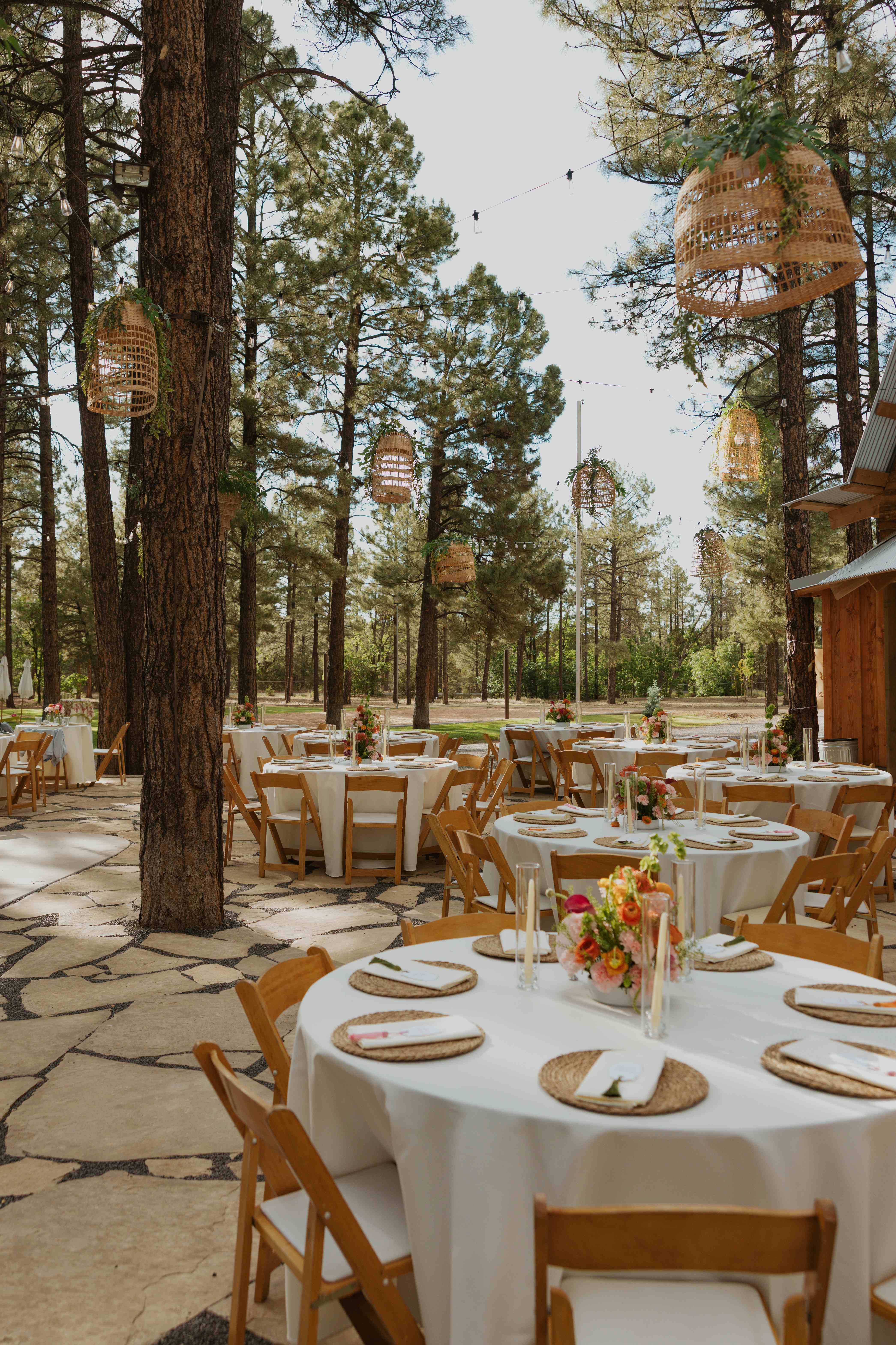 Daytime reception with round tables, woven placemats, floral centerpieces, and taper candles beneath woven pendant lanterns in the pines