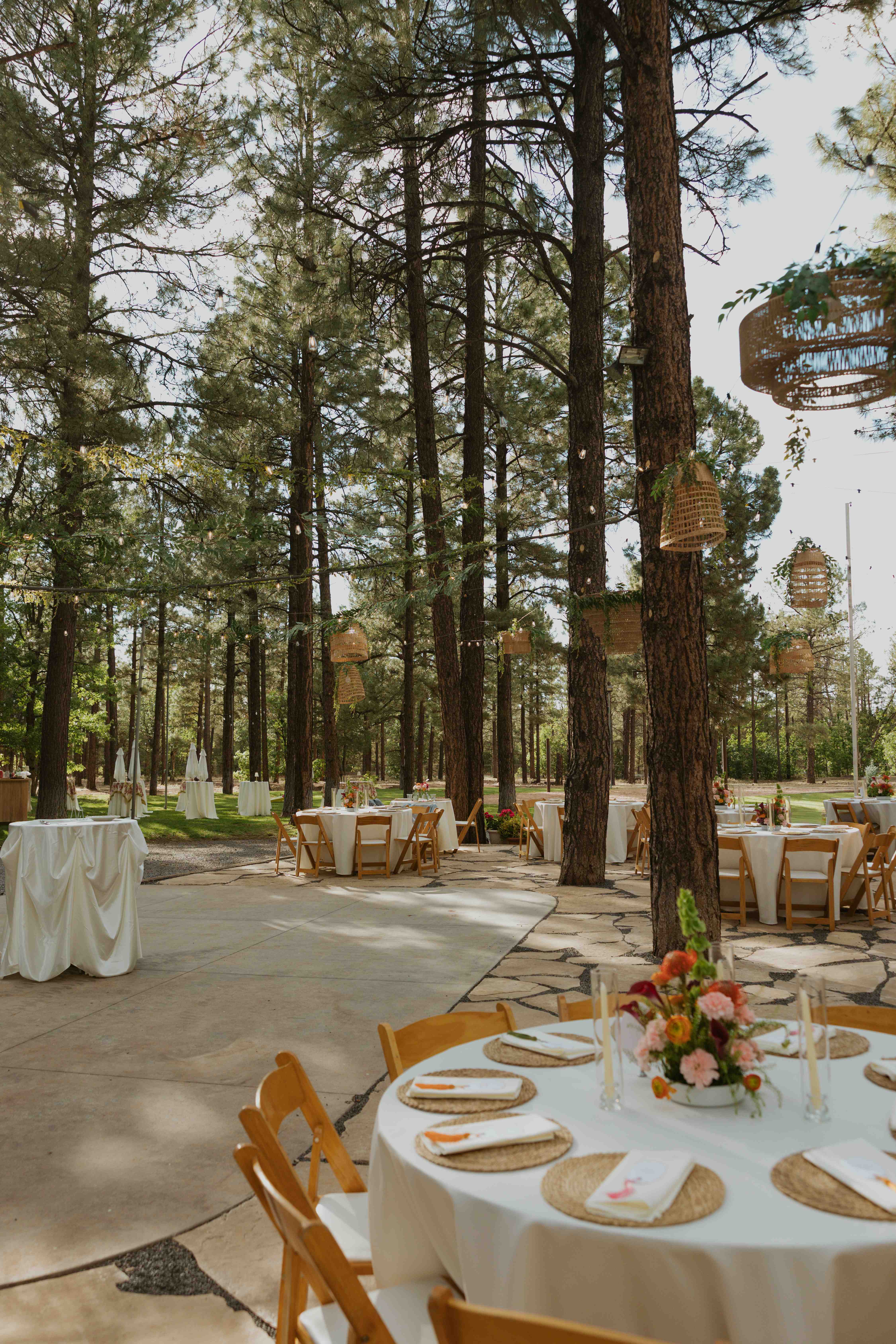 Daytime reception with round tables, woven placemats, and floral centerpieces beneath woven pendant lanterns on a flagstone patio