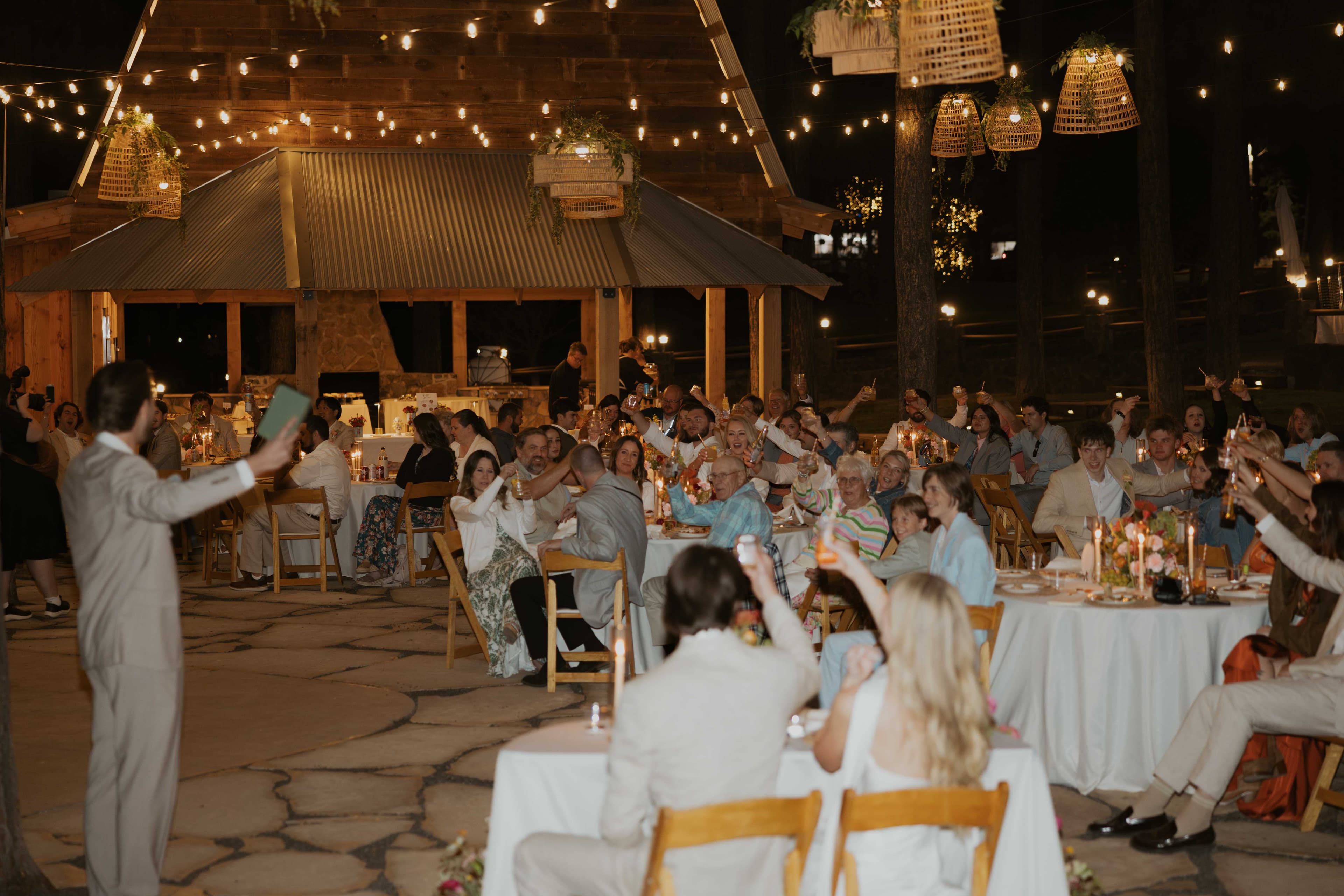 Guests raising their glasses in a toast at warm candlelit reception tables at night with the cabin glowing behind