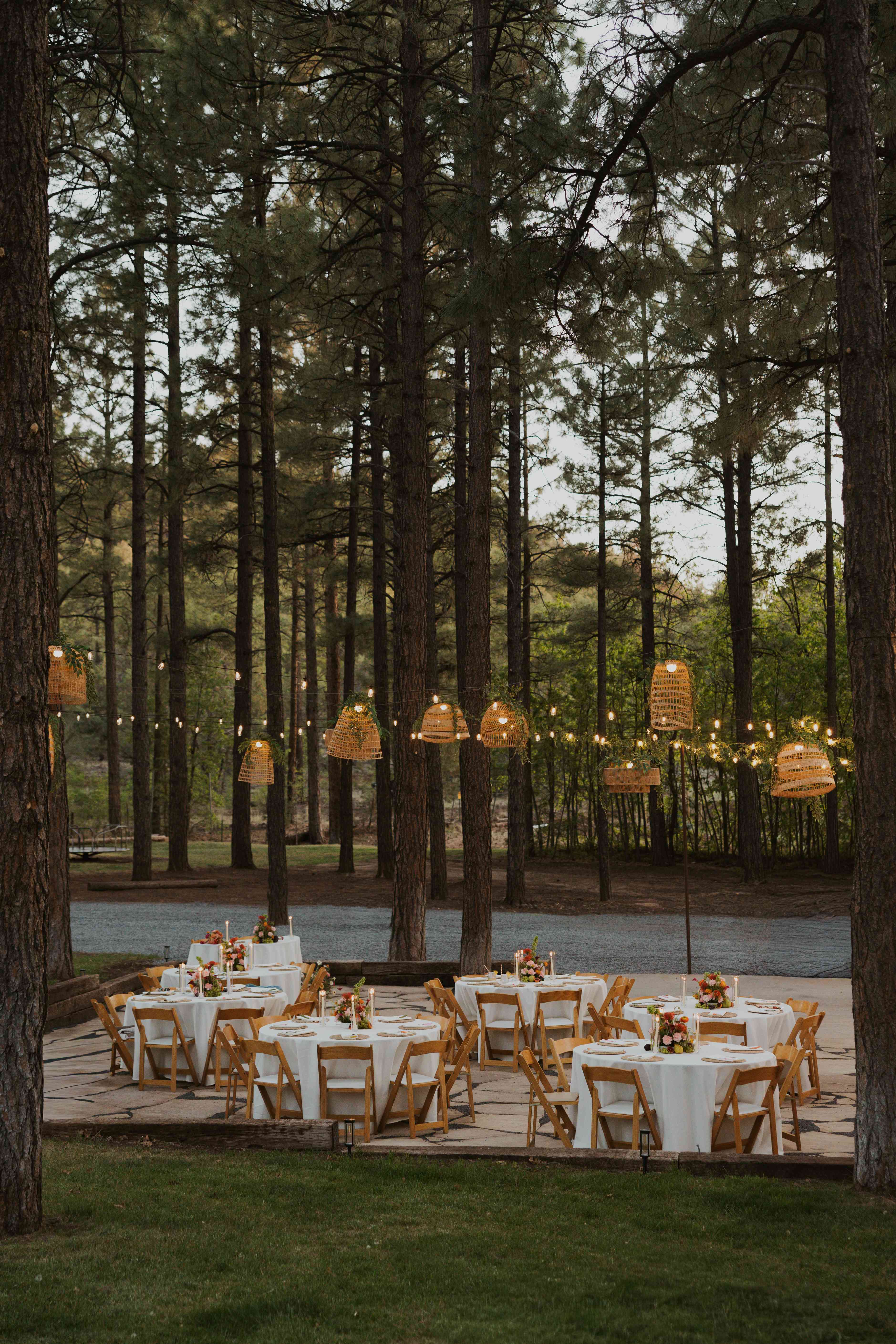 Glowing woven pendant lights and string lights over a flagstone reception with white-linen tables among tall Ponderosa pines at dusk