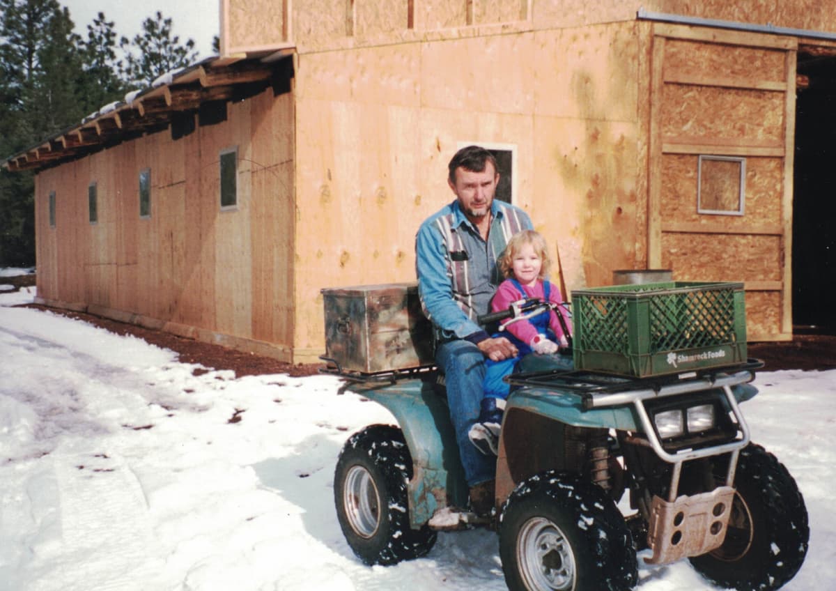 George and a young family member on an ATV in front of a building under construction at the ranch, winter snow on the ground