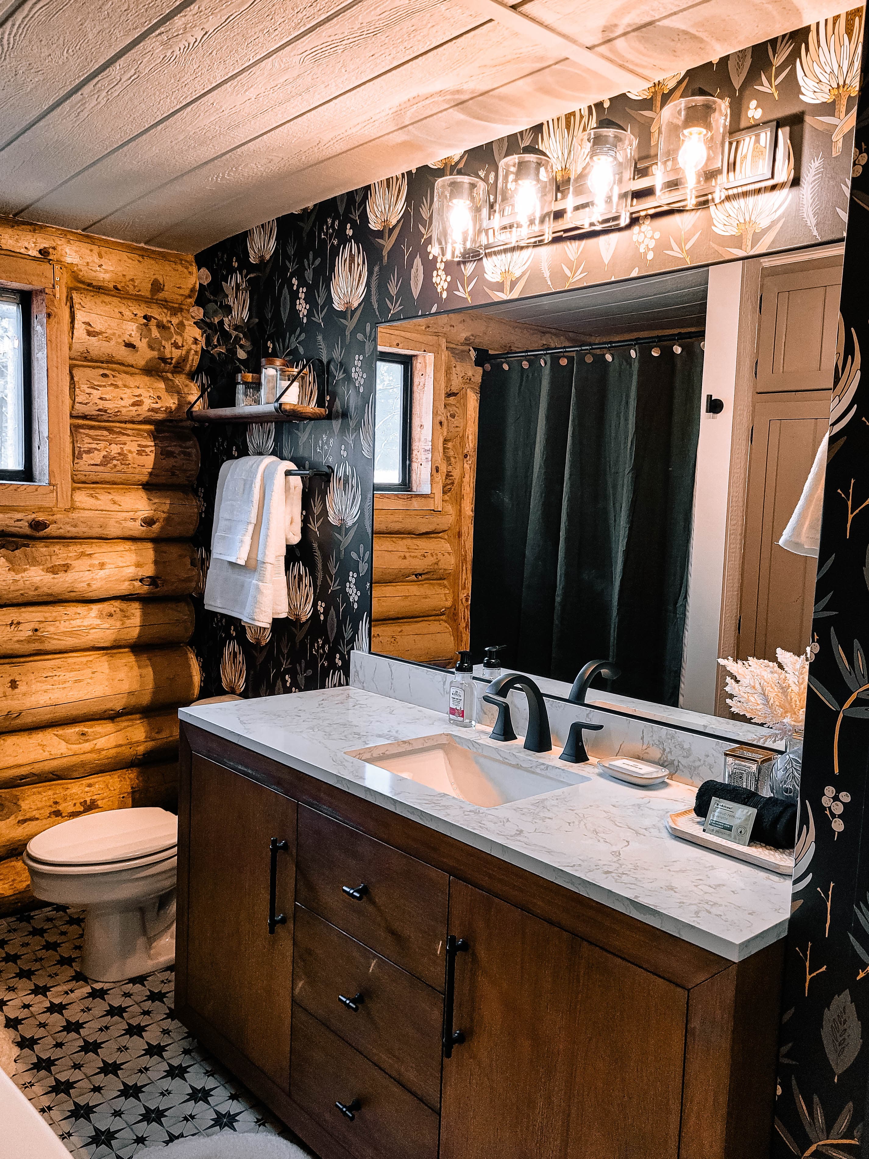 Cabin bathroom with dark floral wallpaper, marble vanity, log walls, black fixtures, and patterned star tile floor