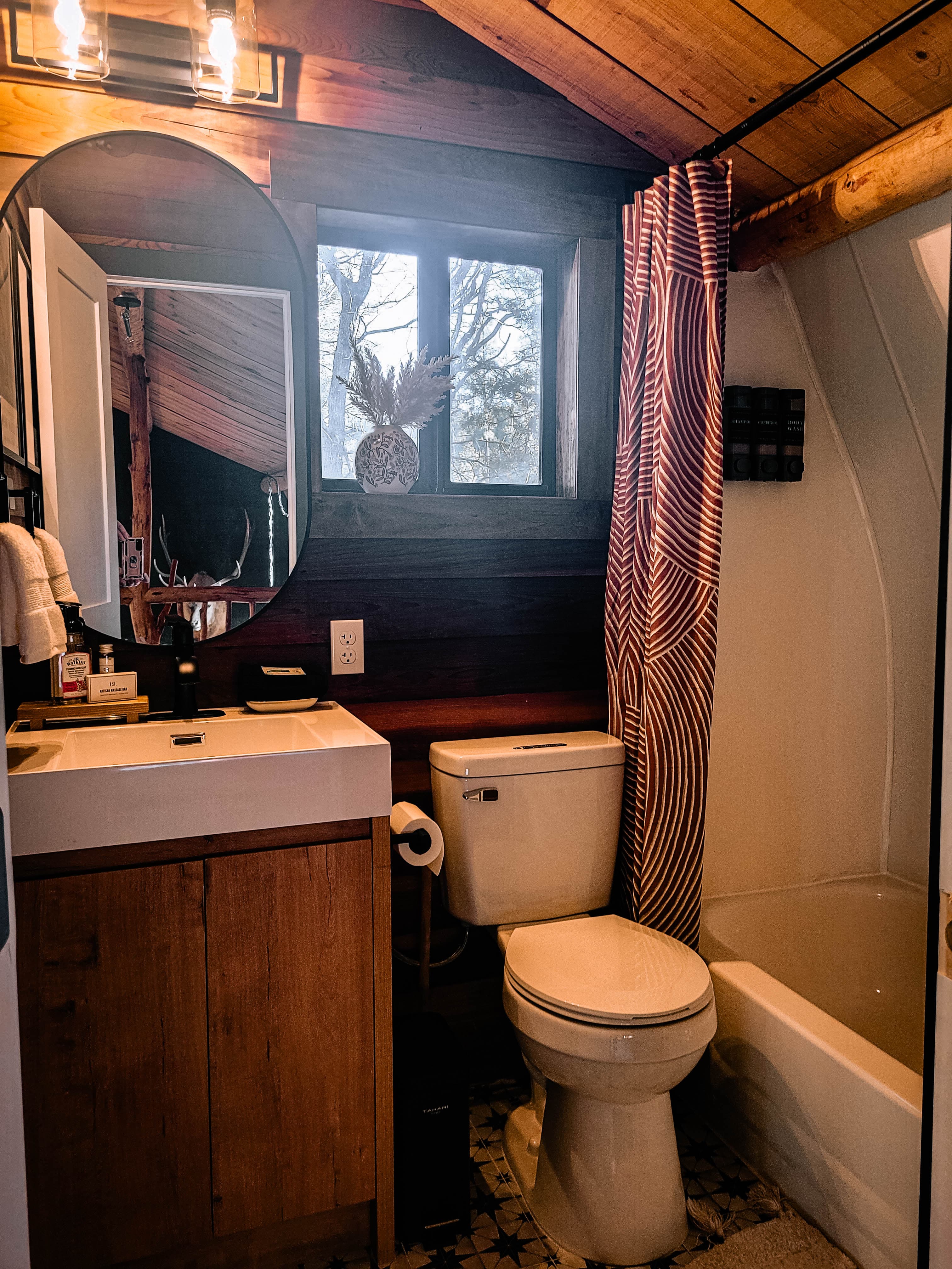 Cabin second bathroom with wood vanity, oval mirror, dark wood accent wall, window with snowy pine view, striped shower curtain over bathtub, and star-pattern tile floor