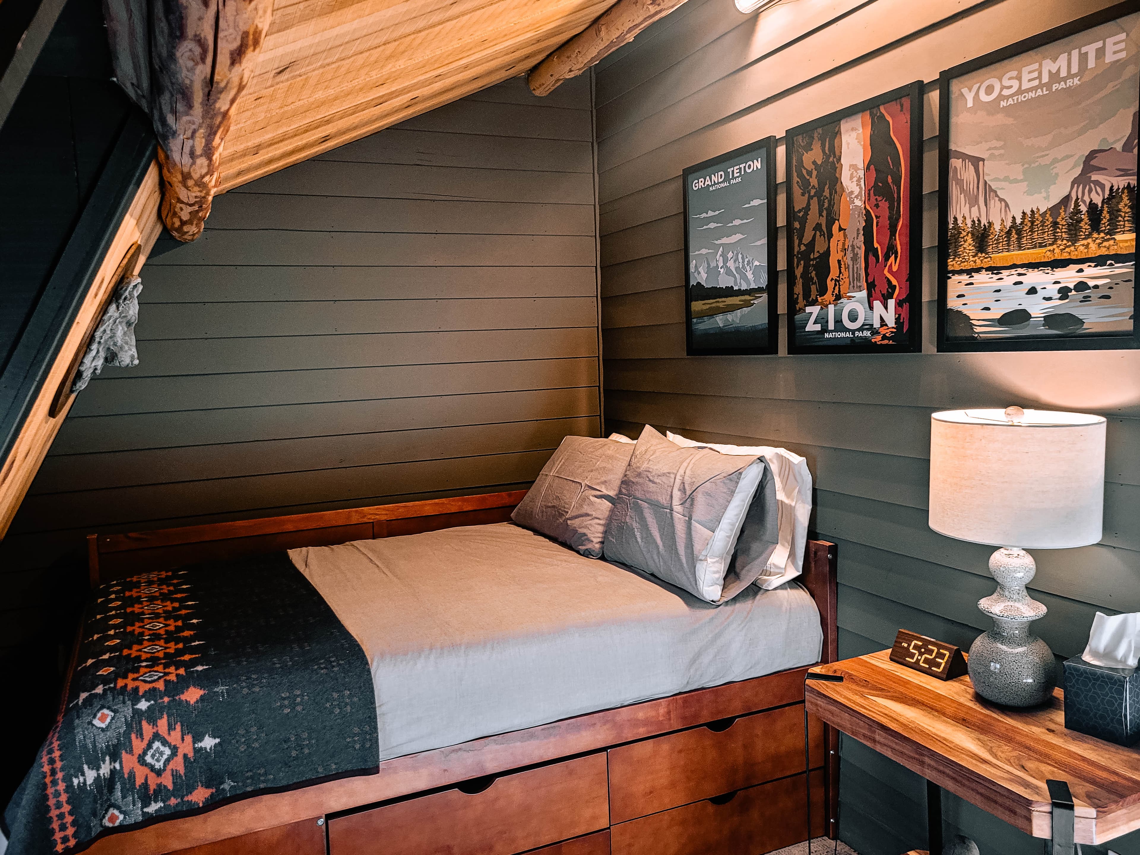 Cabin loft nook with twin bed, Pendleton blanket, angled dormer window, and highland cow and bison medallion wall art