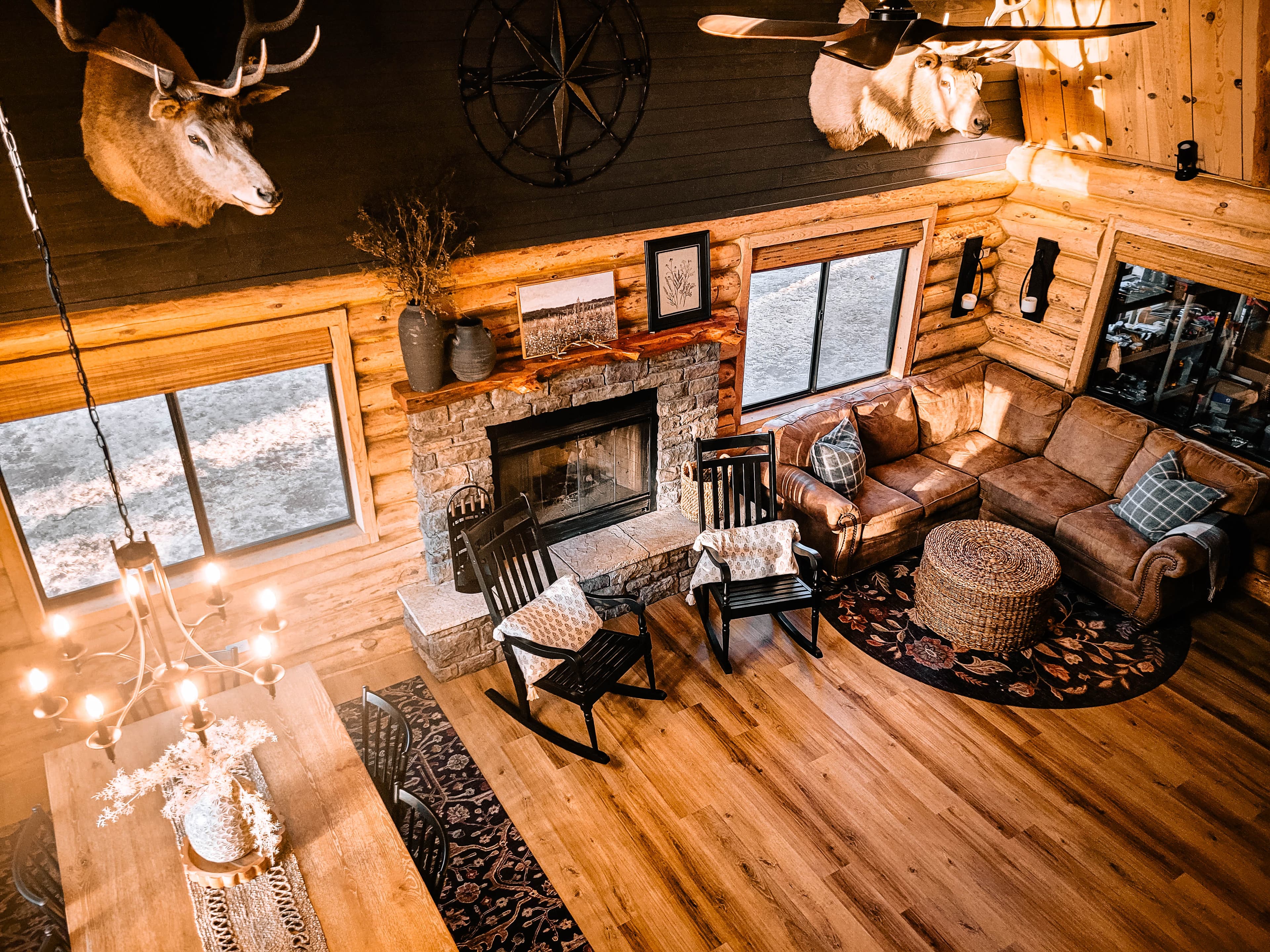 Aerial loft view of the cabin living room showing stone fireplace, dining table with chandelier, and mounted trophy animals