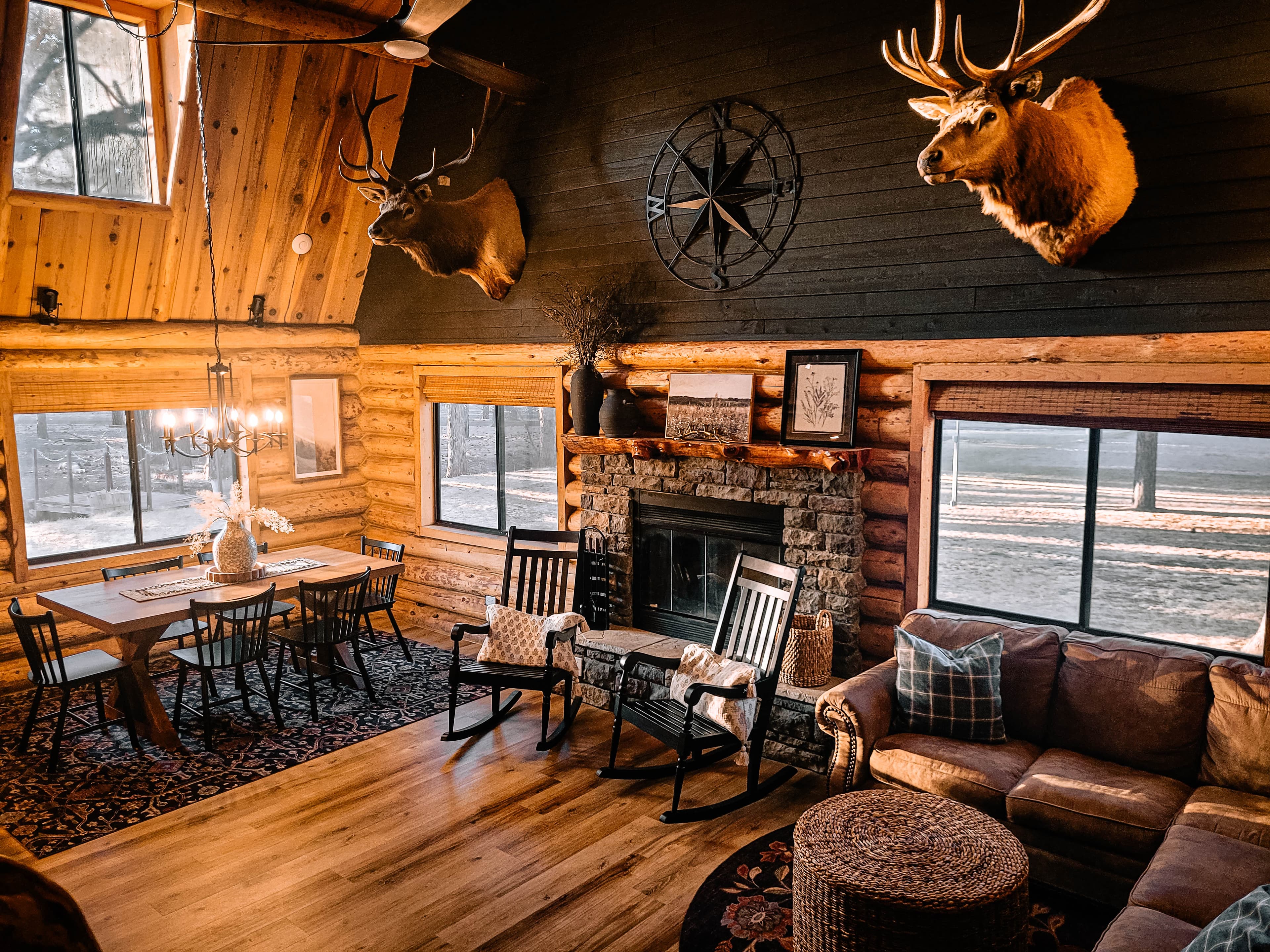 Cabin great room with stone fireplace, leather sectional, rocking chairs, dining table, and mounted elk and sheep heads on log walls