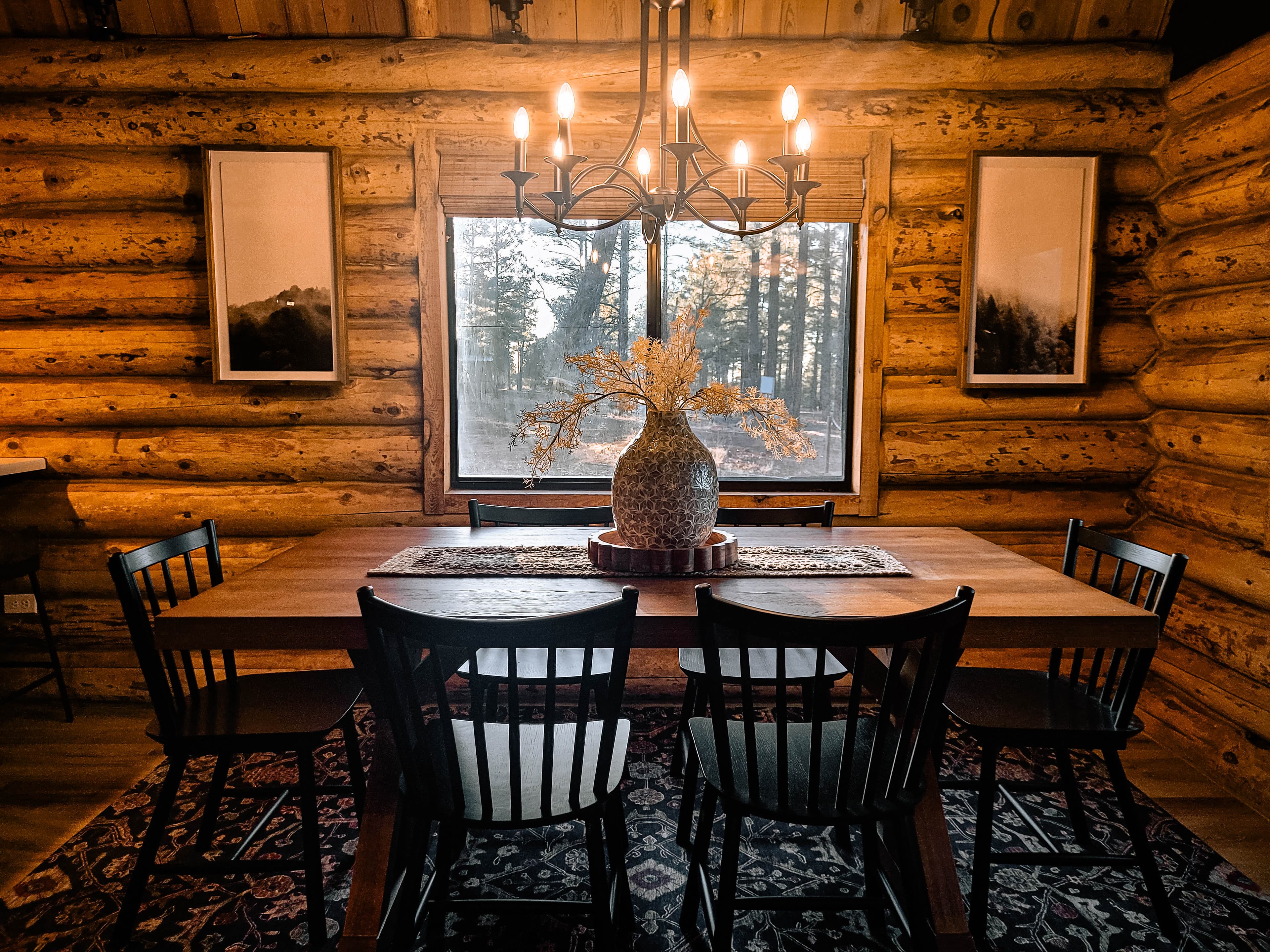 Cabin dining area with farm table, black Windsor chairs, candelabra chandelier, and log walls with window to the pine trees