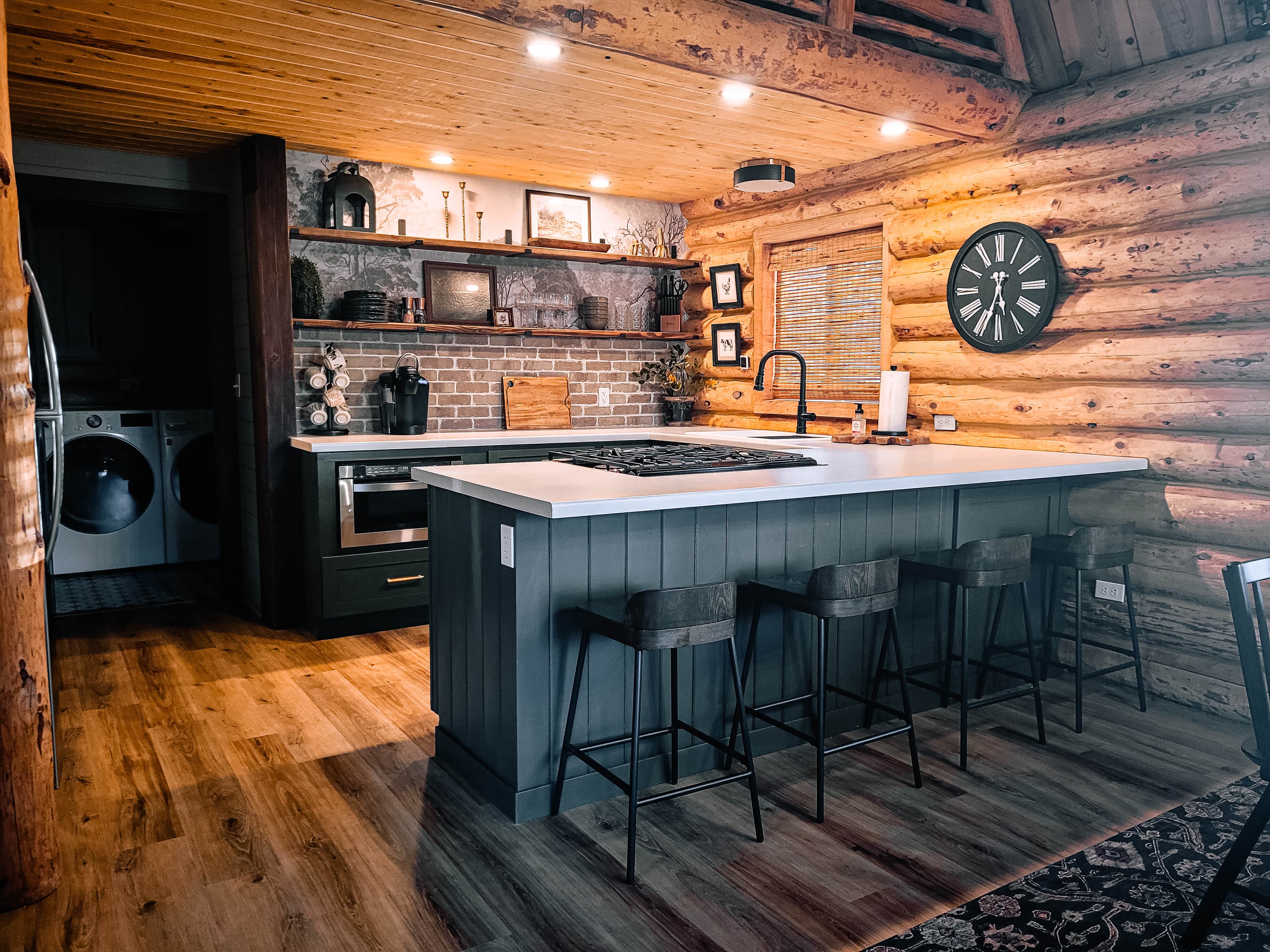 Cabin kitchen with dark teal island, bar stools, brick backsplash, open shelving, gas cooktop, and warm log wall accents