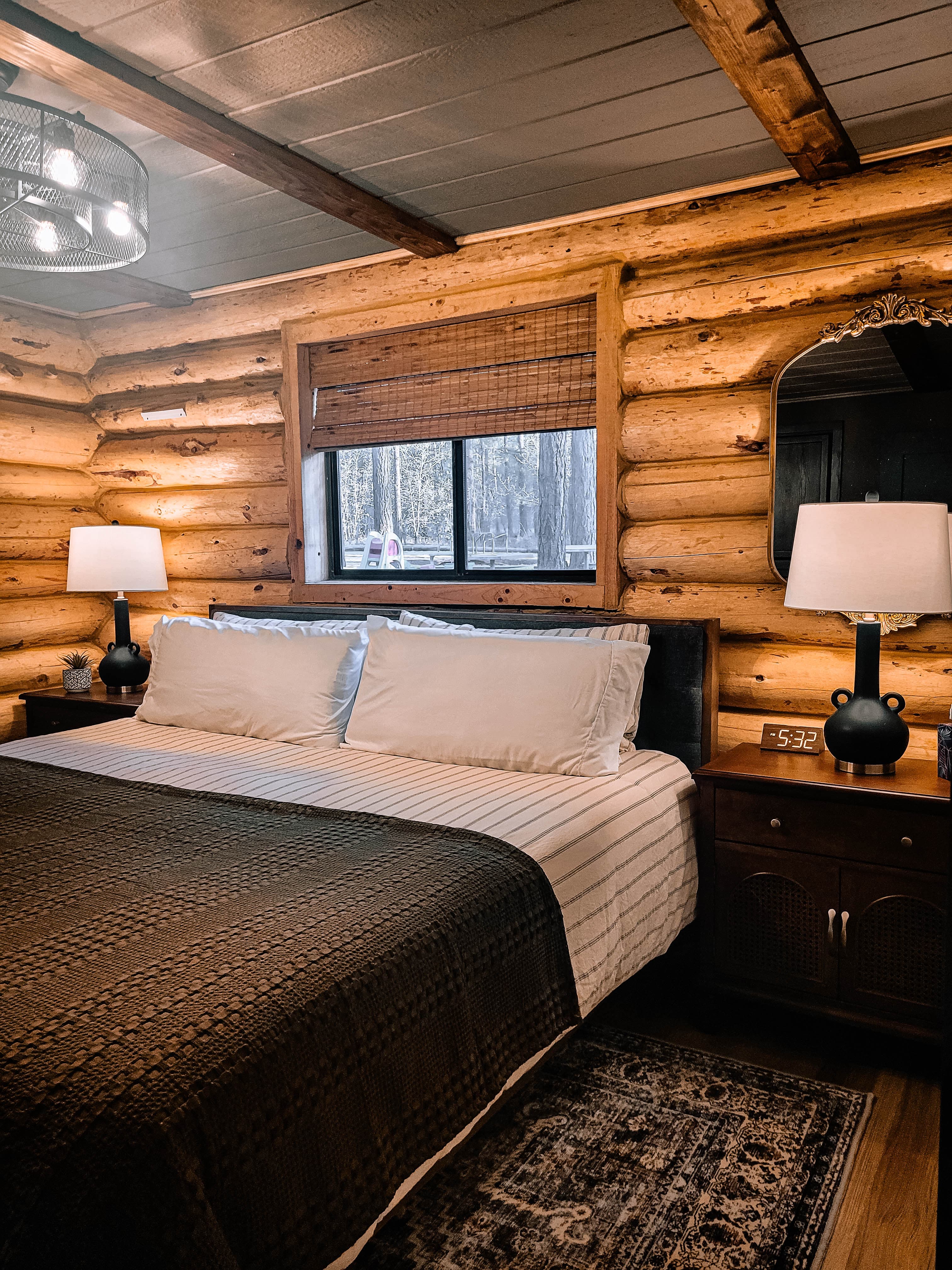 Cabin master bedroom with king bed, dark quilt, log walls, bamboo blinds, and ornate gold mirror