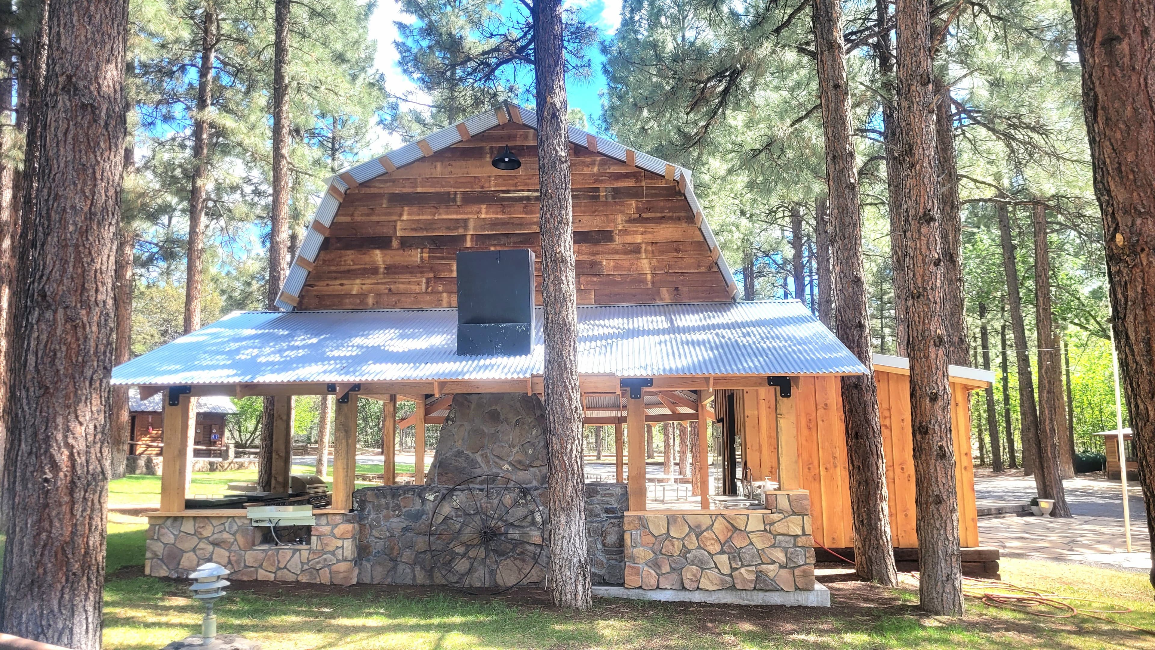 Front exterior of the ramada barn building with round roof, wood siding, and stone chimney through pine trees