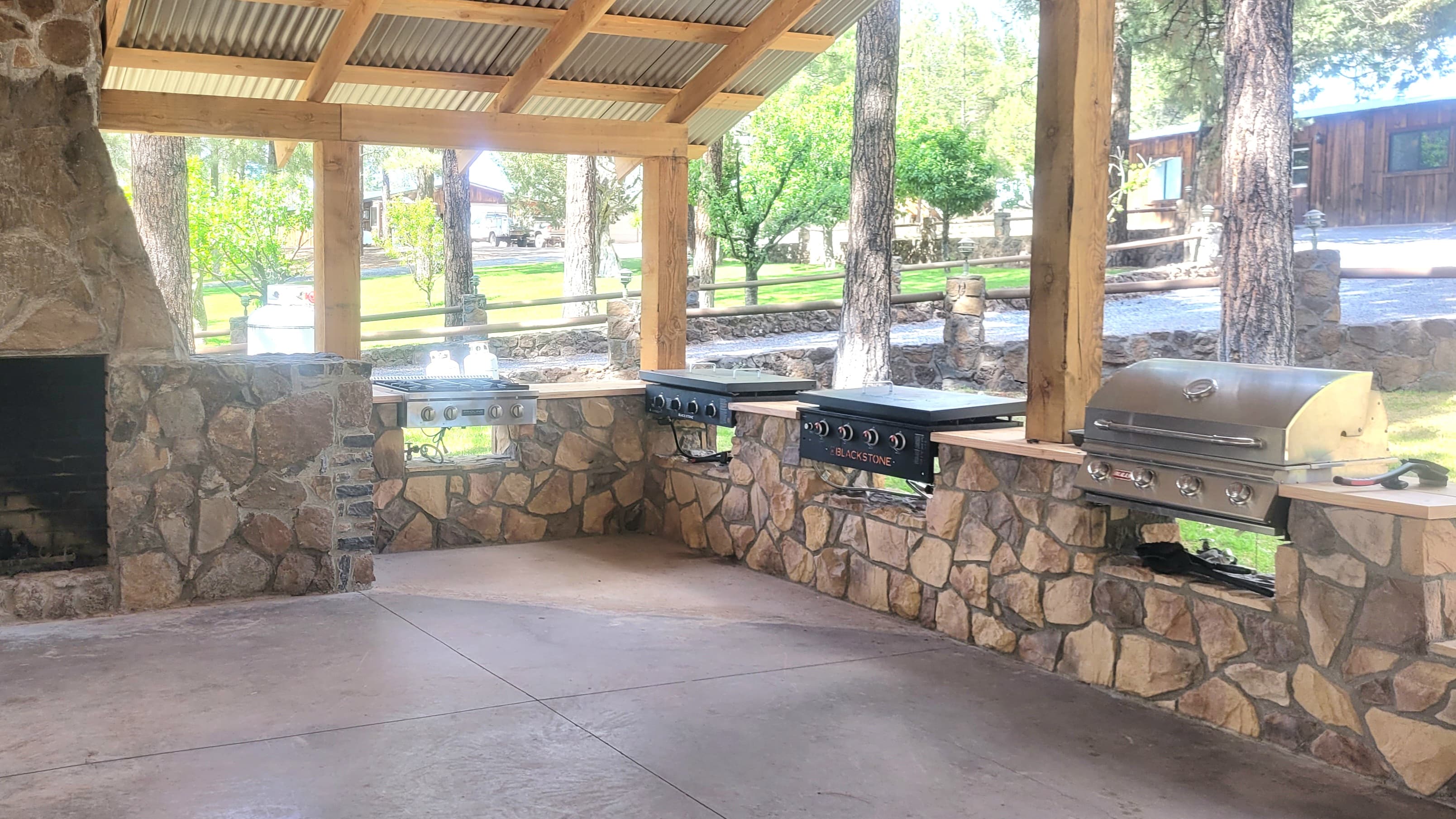 Outdoor BBQ and cooking station with stone masonry counter holding a gas grill, Blackstone flat-top griddle, and gas burner under a corrugated metal roof