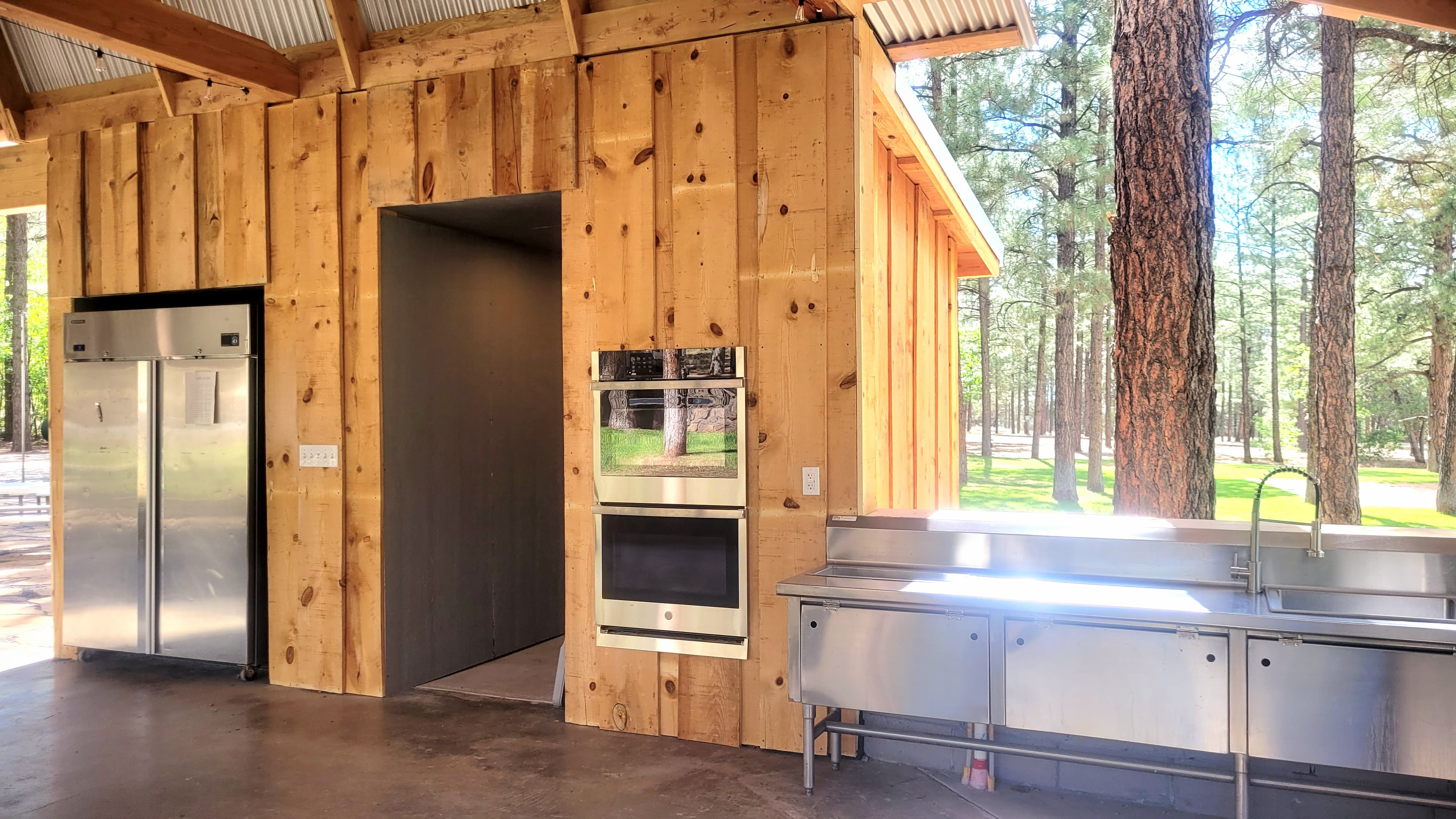 Ramada kitchen interior showing stainless fridge, double wall oven, and stainless prep sink with ponderosa pines visible through open sides