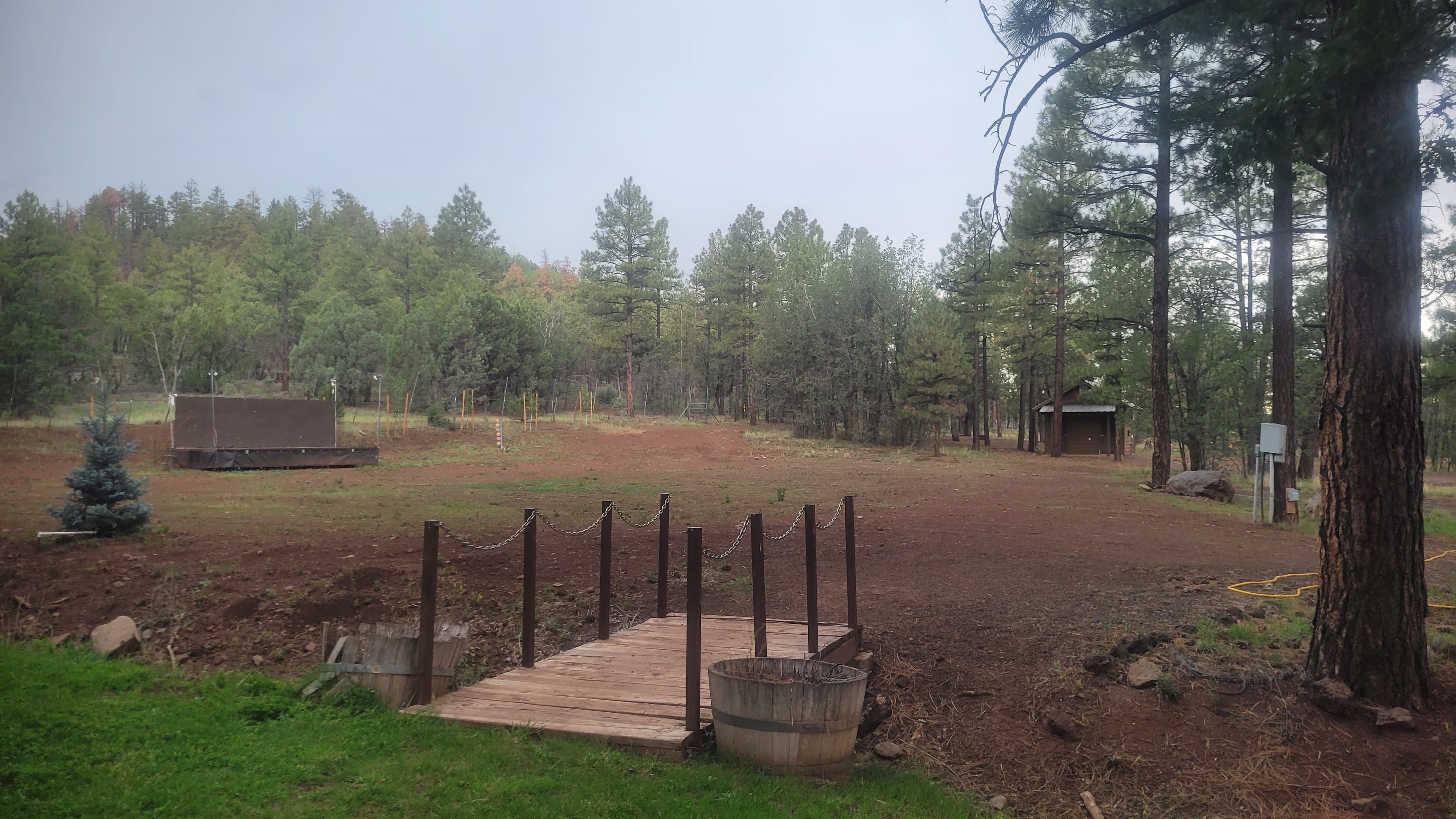 Campground activity area with a small wooden footbridge, chain railing, barrel planters, archery and game area, and a small shed shelter among ponderosa pines