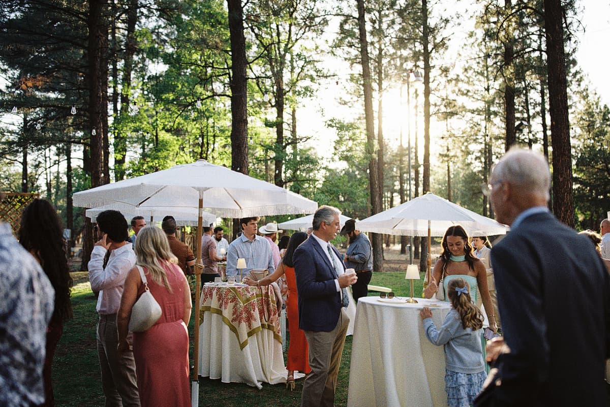 Guests mingling at a golden-hour outdoor cocktail reception at Diamond W Ranch, under white fringed market umbrellas among tall ponderosa pines with string lights overhead